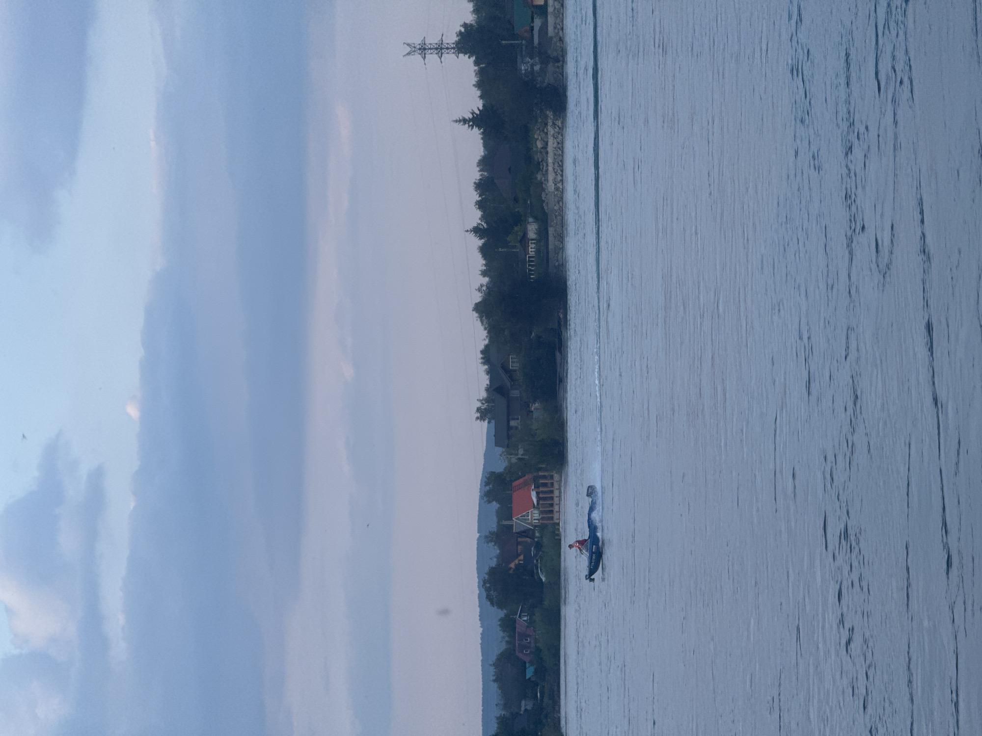 In Irkutsk, Russia, on a cloudy day, a man in a red shirt pilots a small, dark-colored motorboat across a wide, calm body of water. The boat is moving at speed, evidenced by the spray trailing behind it. In the background, a shoreline is visible, lined with trees and houses, some with red roofs. A tall electricity pylon stands out against the skyline, and rolling hills form the distant backdrop. The sky is filled with soft, grey and pale blue clouds.