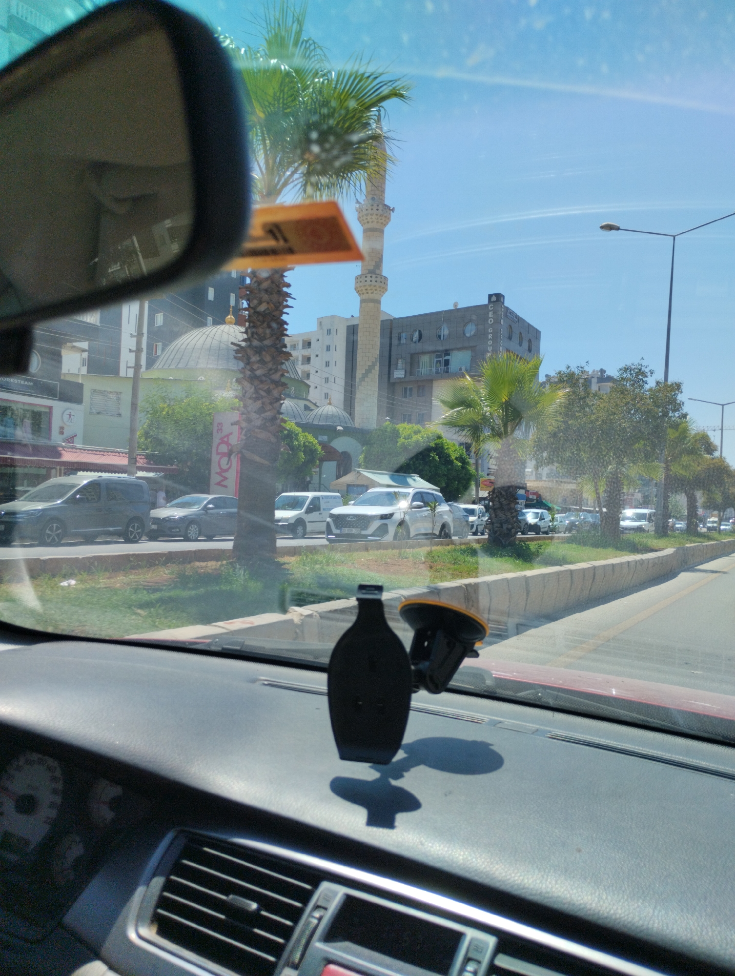The image is taken from inside a vehicle, looking out through the windshield and side mirror. The scene is an urban environment in Mezitli, Turkey, under a clear blue sky.
In the foreground, a black car phone holder with a suction cup is mounted on the dashboard. Part of the dashboard, including an air vent and a portion of the instrument cluster, is visible.
Outside the car, a street scene unfolds. Several vehicles are parked and moving on a road lined with a low concrete barrier and green grass. Palm trees and other foliage are present.
In the background, there are multi-story buildings. One building features a distinctive dome and a minaret, suggesting a mosque. Another building has circular window features and the text "CEO GROUP" is visible. A street lamp stands tall on the right side of the road. The overall impression is of a sunny day in a coastal Turkish city.