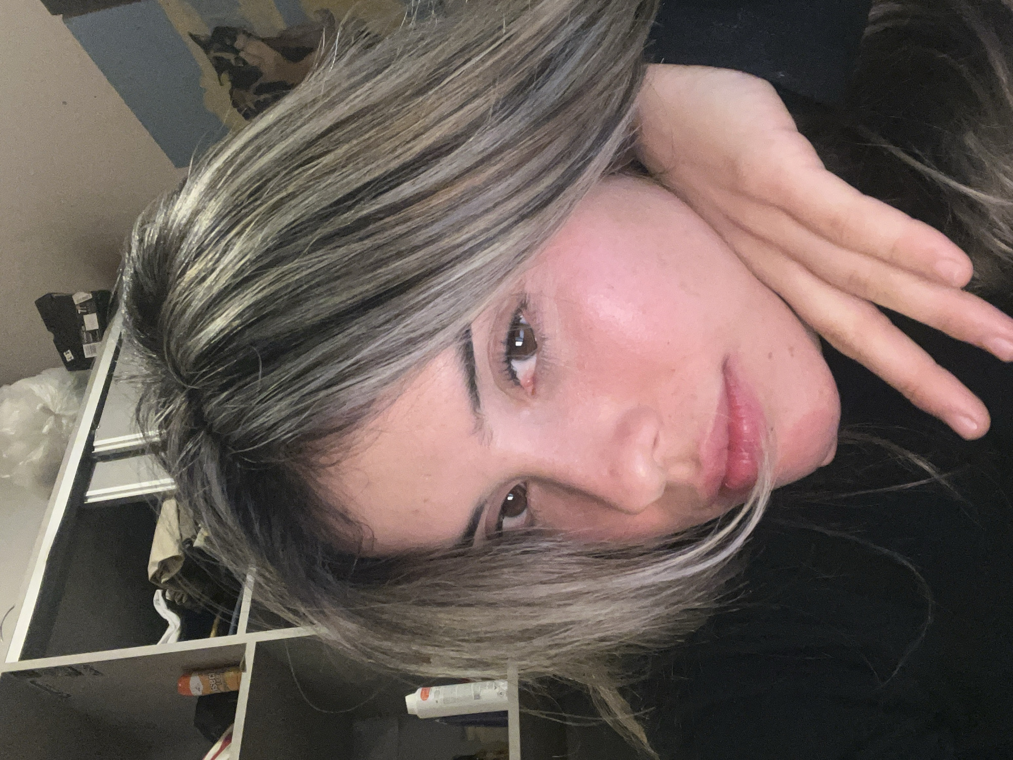 A young woman with layered blonde and brown hair is shown in a close-up, selfie-style shot. She is wearing a black garment and her left hand is resting on her chin, with her fingers extended. Her head is tilted, and her gaze is directed towards the camera. The background appears to be an indoor setting, with shelves visible on the left side of the image containing various items, including bottles and what looks like a bag of trash. A portion of a blue and yellow striped wall is visible on the right. The lighting is dim, creating shadows and highlighting the woman's features. The image does not contain any discernible elements that would indicate it was taken in Sumalao, Argentina.