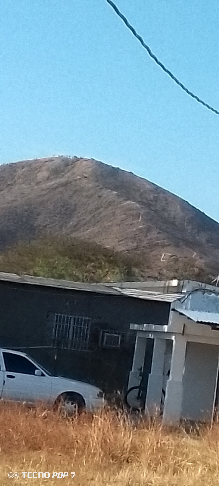 A photograph taken in Altagracia, Venezuela shows a dry, brown-toned mountain under a clear blue sky. A power line cuts across the upper-right corner of the frame. In the foreground, a white sedan is parked in dry, yellow grass. Behind the car is a dark-colored building with barred windows and an air conditioning unit visible. A white-columned structure is to the right of the building, partially obscuring a bicycle. The overall scene suggests a rural or semi-rural environment.
