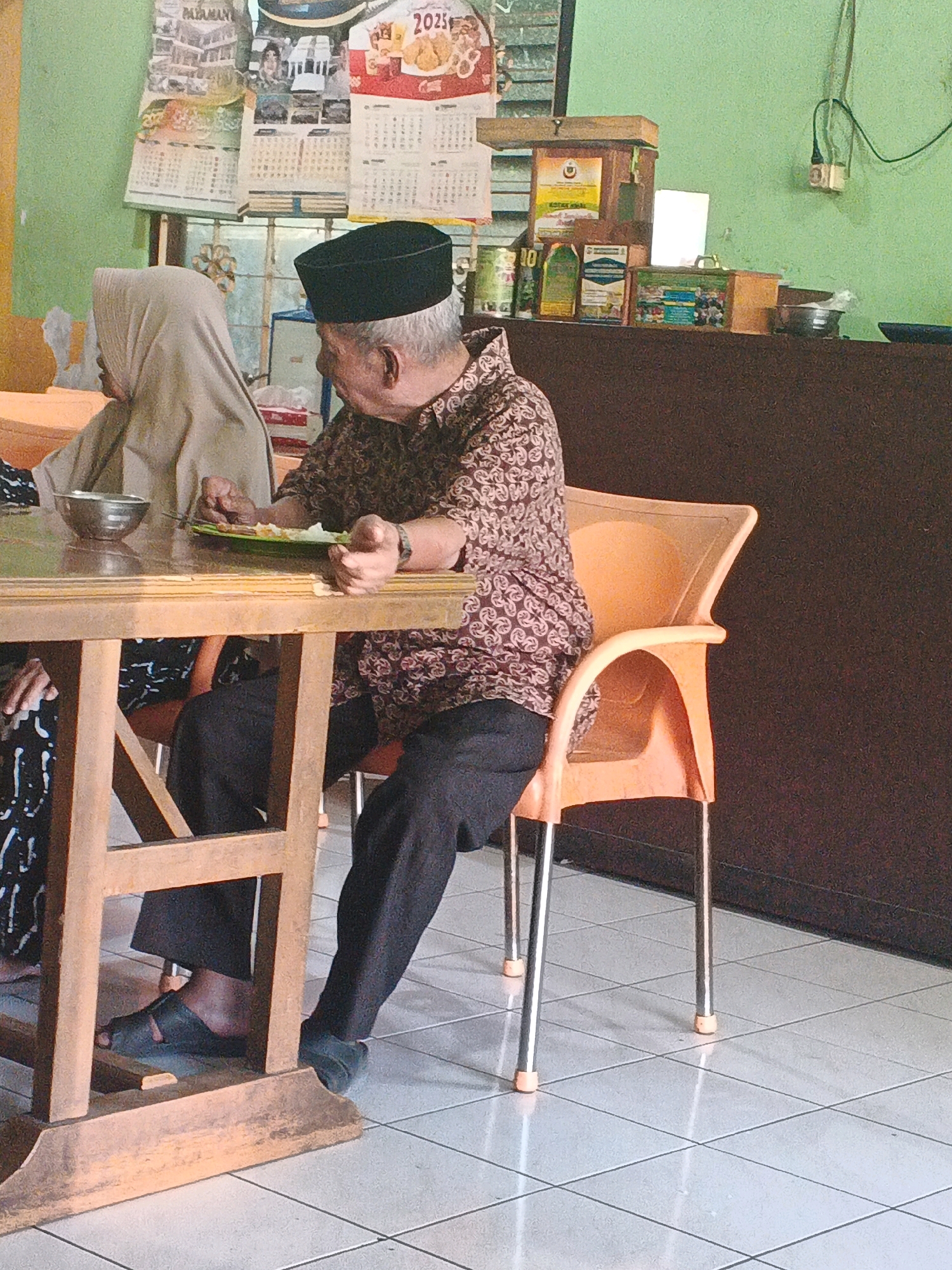The image captures an indoor scene, likely a casual dining establishment or home in Ungaran, Indonesia. In the foreground, an elderly man wearing a patterned brown shirt and a black peci (cap) is seated at a wooden table. He is holding a fork and a plate with food, appearing to be in the process of eating. To his left, a woman wearing a beige hijab is also seated at the table, partially visible. The table is made of wood and has a light-colored surface. In the background, a dark brown counter is visible, holding various items including containers, cans, and what appears to be a computer monitor. Above the counter, calendars are displayed on a light green wall. One calendar clearly shows "2025". The floor is tiled with a light grey pattern. An orange plastic chair is positioned next to the man. The overall lighting suggests an indoor environment with natural light coming from the side.