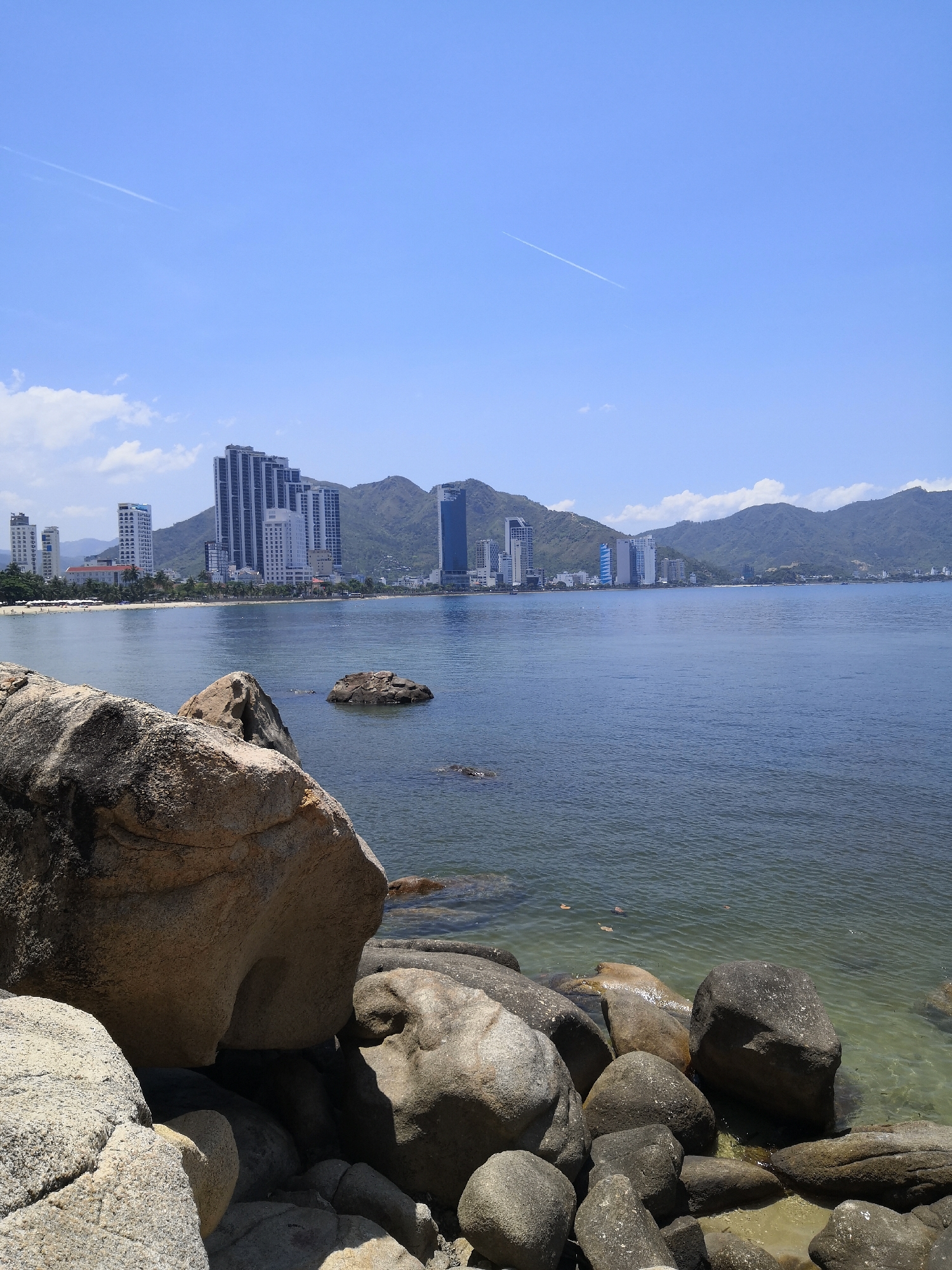 This image depicts a coastal scene in Bac Nha Trang Ward, Vietnam, under clear skies. In the immediate foreground, large, weathered granite boulders of varying sizes and tones, from light tan to dark grey, occupy the lower left and right sections. The water directly adjacent to these rocks is clear, revealing the seabed and submerged stones. Further into the midground, the sea exhibits a calm, dark blue-green hue with subtle surface ripples. Several isolated, dark rocks protrude from the water. Three light-colored, possibly organic, objects float on the water's surface in the middle-right.
Across the bay, a long sandy beach is visible, populated by numerous distant, indistinct figures. Behind the beach, a dense urban skyline features a concentration of high-rise buildings, including numerous modern skyscrapers and hotels, primarily in shades of white and grey. These structures are nestled against a backdrop of undulating, green-covered mountains that extend across the horizon. The sky is bright blue with scattered white cumulus clouds, predominantly on the left side and above the mountains. A faint white aircraft contrail diagonally traverses the upper left quadrant of the sky. The scene is illuminated by strong sunlight, indicating a clear day. No specific actions or interactions of individuals are discernible.