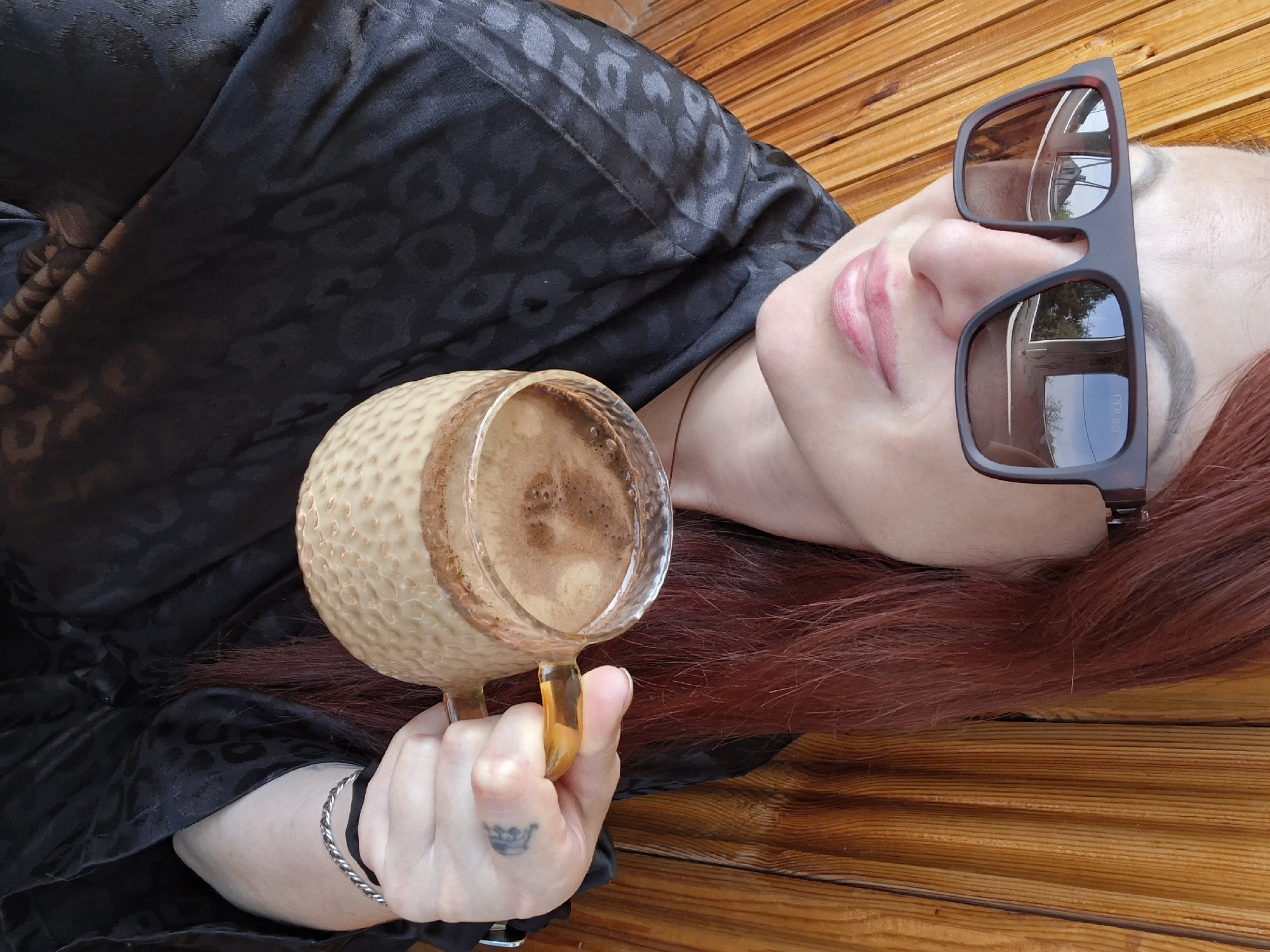 A medium shot photograph shows a woman with reddish-brown hair, wearing large, dark sunglasses and a black patterned robe, holding a mug of coffee. She is positioned in front of a wooden paneled wall. The sunglasses reflect a building and trees. Her lips are slightly parted, and she has a small tattoo on her right hand. The coffee in the mug is frothy. The background appears to be an outdoor setting with a rustic wooden wall. The location is the Забалоцкі сельскі Савет in Belarus.