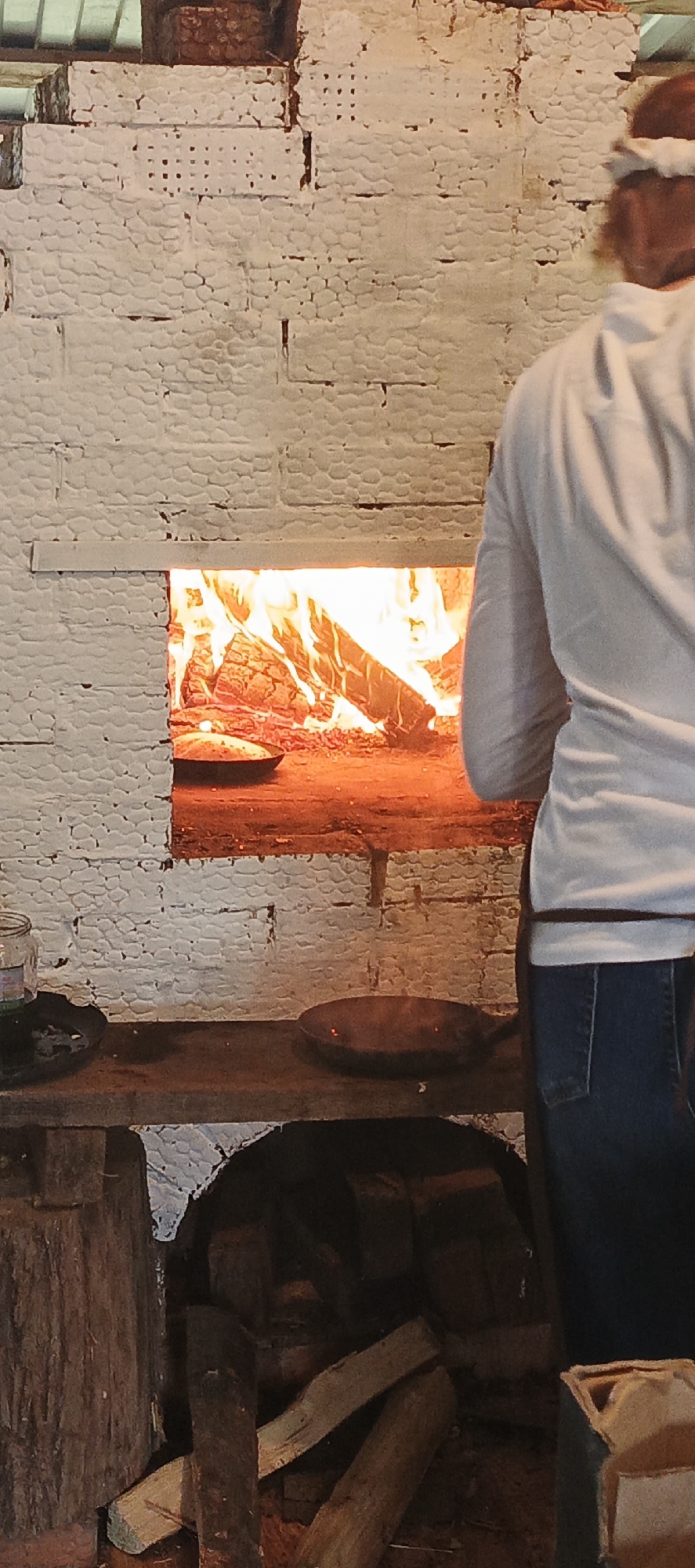 The image depicts a person, viewed from the back, standing in front of a rustic, white brick oven with a lit fire inside. The oven opening reveals burning logs and a metal dish, possibly containing food, placed on the oven floor. The person is wearing a white long-sleeved shirt and blue jeans, with an apron tied around their waist. To the left of the oven, a wooden shelf holds a glass jar and another metal dish with a faint red glow. Below the shelf, a collection of firewood is visible within the lower compartment of the oven structure. The overall scene suggests a traditional cooking or baking setting, potentially in a rural or historical context, given the materials and methods employed. While the prompt mentions Старые Быги, Russia, this specific location cannot be visually confirmed from the image alone, but the architectural style and materials are consistent with elements found in that region.