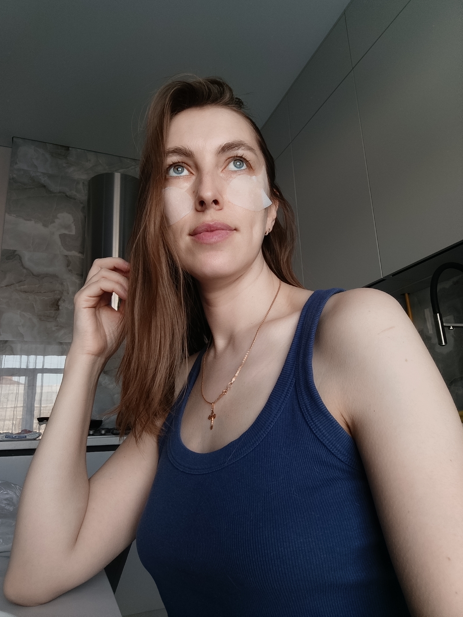 A young adult Caucasian woman with long, light brown hair, blue eyes, and fair skin is seen from a medium-angle, low-angle perspective. She is wearing a dark blue ribbed tank top and a gold chain necklace with a cross pendant. Under-eye masks are applied to her face. Her left hand is raised, lightly touching her hair. The background reveals a domestic kitchen setting with marble-patterned wall panels, a stainless-steel range hood, and a black faucet. The photo appears to have been taken in a residential area of Chaikovsky, Russia.