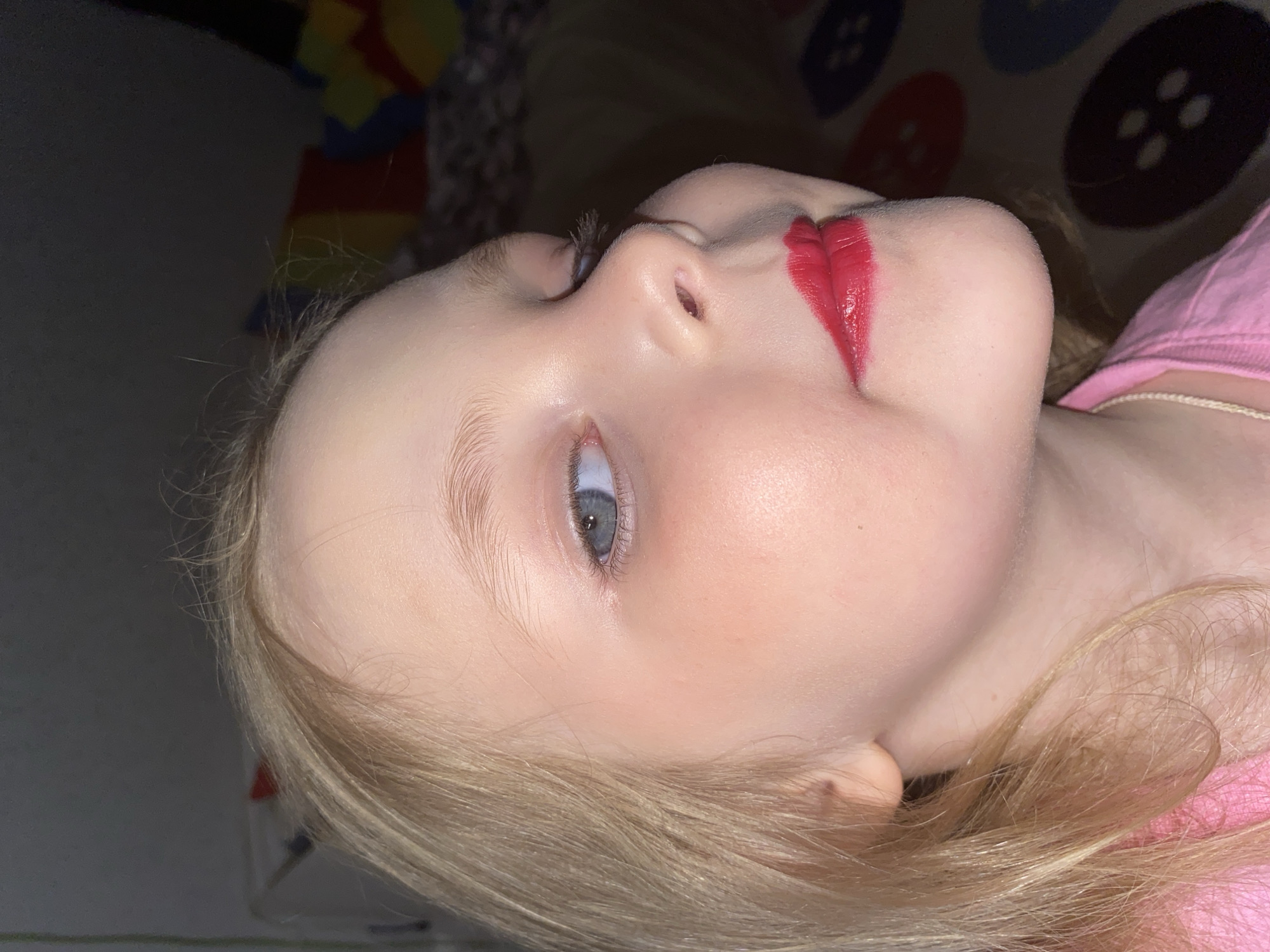 A close-up, portrait-oriented photograph, rotated 90 degrees counter-clockwise, shows a young child with fair skin and light blonde hair. The child is depicted from the chest up, lying down in an indoor setting in Votkinsk, Russia, with their head tilted upwards and eyes looking slightly to the viewer's left.
The most striking feature is the child's mouth, which is generously covered in bright red lipstick, applied beyond the natural lip line, suggesting playful experimentation with makeup. Their eyes are a clear blue-grey and are wide open, conveying a neutral or subtly curious expression. Fine, light blonde hair frames the child's face and falls around their shoulders. They are wearing a pink top or t-shirt, visible on their right shoulder and upper chest.
The background features patterned fabric, likely a blanket or bedding. To the left of the child's head, there are colorful geometric shapes, including yellow and red. To the right, a darker fabric displays large circular designs resembling buttons, in various colors such as dark blue, black, red, and light blue. The lighting is soft and appears to be from an indoor source. No text is visible in the image.