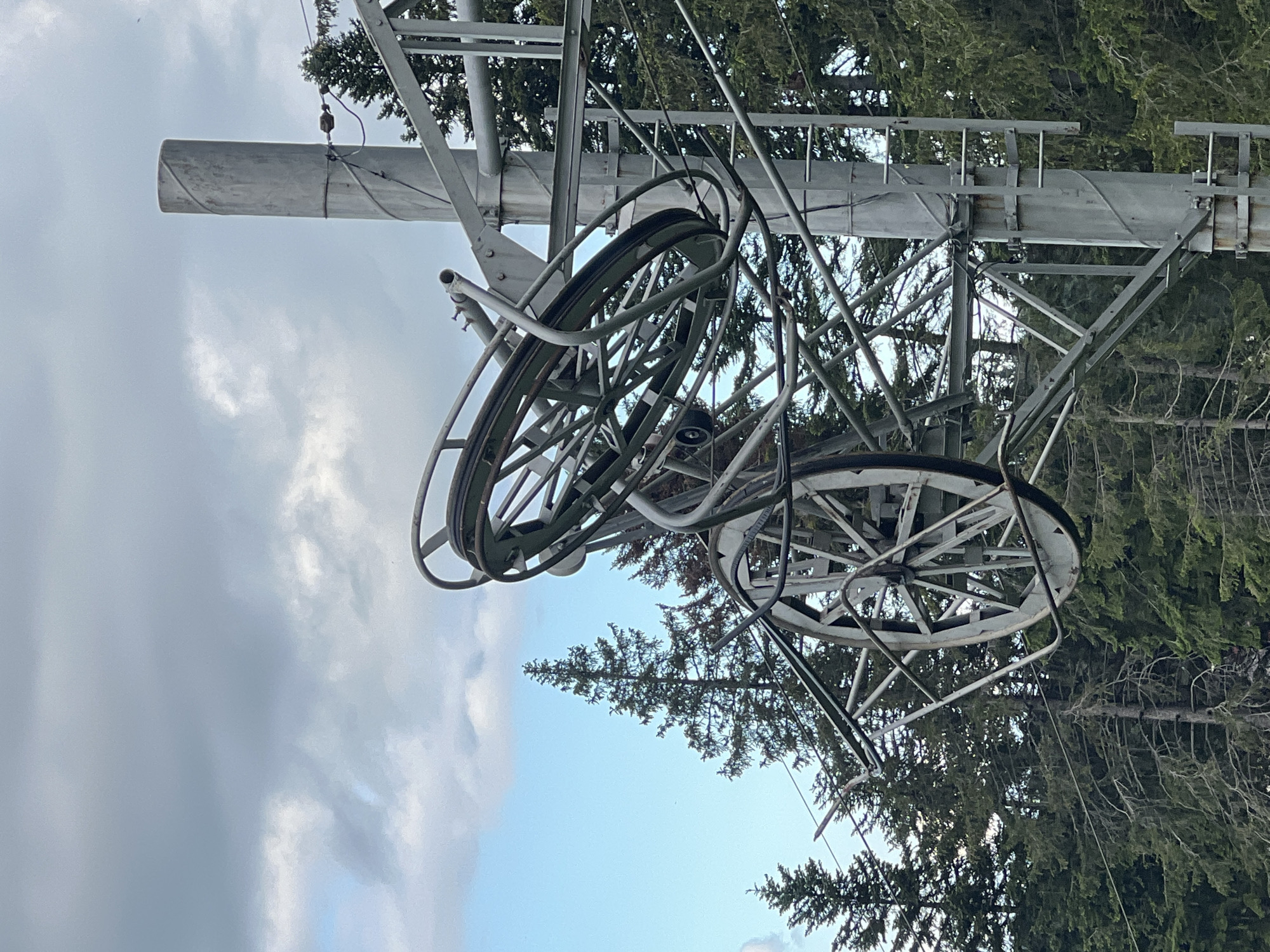A ski lift tower and pulley system are visible against a cloudy sky and a backdrop of evergreen trees. The tower is a large, metallic structure, supporting the complex arrangement of pulleys and cables that form part of the ski lift mechanism. The pulleys themselves are large, circular metal wheels, with some showing signs of wear and rust. Cables are strung between different points of the structure, disappearing into the surrounding trees. The trees are dense and tall, typical of a mountainous or forested environment. The overall impression is one of industrial infrastructure within a natural setting.