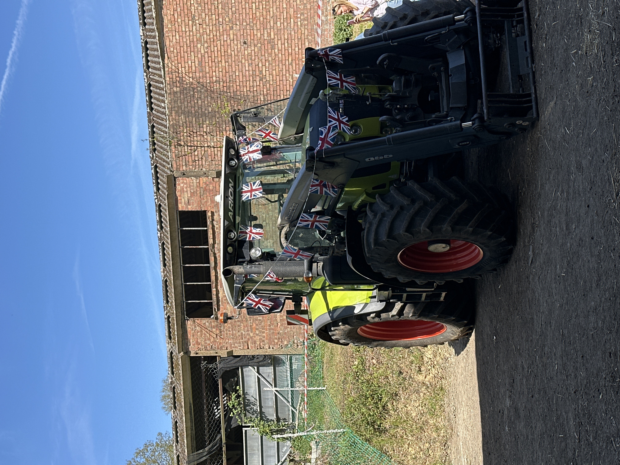 The image captures a green and black Claas Arion tractor, adorned with British Union Jack flags, parked on a patch of dirt and short grass. Behind the tractor is a dilapidated brick structure with missing windows and exposed metal beams, partially covered with chain-link fencing and vegetation. A clear blue sky with a faint contrail is visible overhead. The scene is set in what appears to be a rural or semi-rural area of Sevenoaks, United Kingdom. The photo appears to have been taken at an odd angle and edited to be vertical.