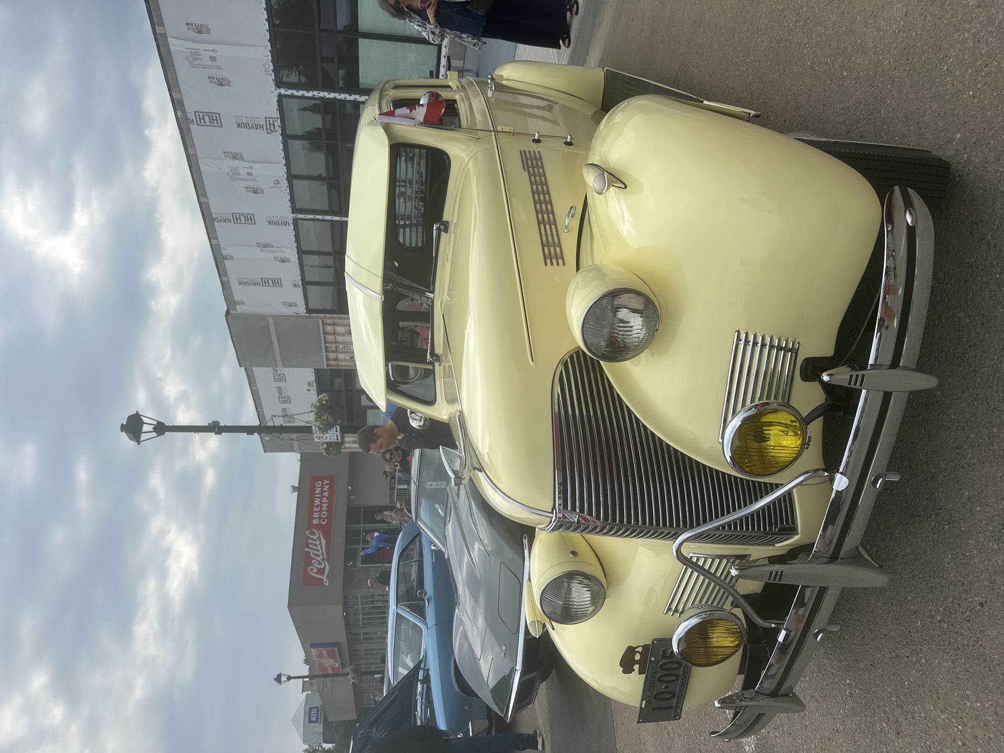 This media file captures a portion of a car show, likely in Leduc, Alberta, Canada, based on the visible signage. The main subject is a light yellow vintage car, prominently displayed at the forefront. Its chrome details, including the grille, headlights, and bumper, gleam. The car is adorned with a small Canadian flag on its driver's side.
Behind the vintage car, other vehicles are visible, including a dark gray car and a blue car, suggesting a line-up typical of car shows. In the background, a building with "Leduc Brewing Company" signage is clearly visible, indicating the location. The building appears to be under some construction or renovation, with materials like "TITAN" and "HAYDUK HARDWARE" visible on sheeting covering parts of the facade.
Several people are scattered around the scene, some looking at the cars, others conversing. The overall atmosphere appears to be casual and leisurely, fitting for an outdoor event. The sky is cloudy, suggesting overcast weather, and the lighting indicates it is likely daytime. The presence of streetlights and the type of buildings imply an urban or town setting. A license plate on the yellow car reads "ALBERTA 10-005 EXPIRES MARCH," providing specific location and temporal context.