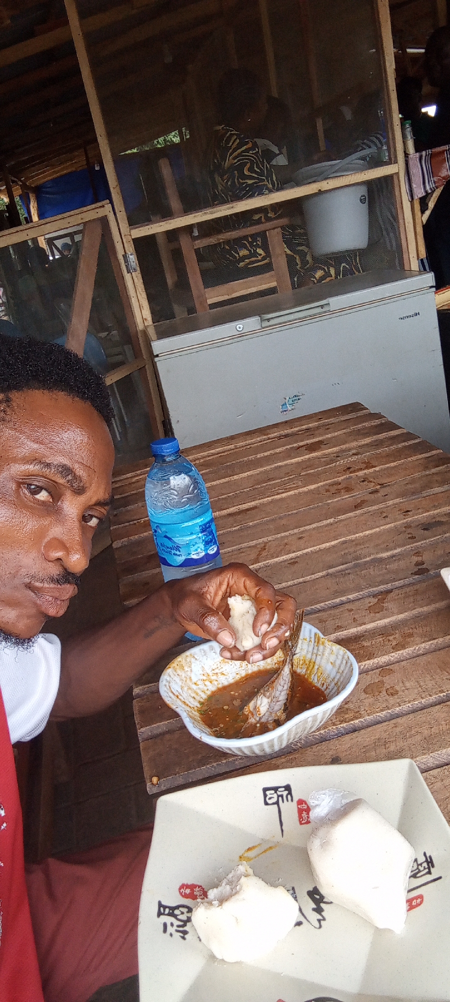 A man is eating a meal at a table in Unknown, Nigeria. He holds a piece of white food in his right hand over a bowl containing a fish in a brown sauce. On a plate next to the bowl are two more pieces of the same white food. A bottle of water is on the table. Behind the table is a building constructed with wooden beams and wire mesh. Inside the building, a white bucket hangs from the ceiling. A white freezer stands against the wall.