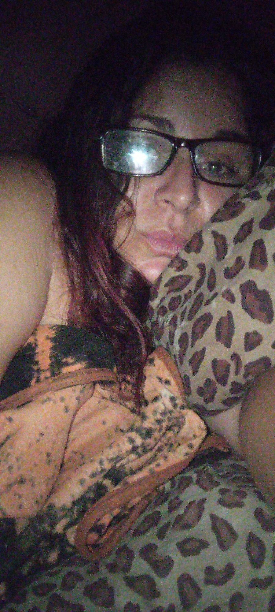 A woman with shoulder-length curly hair and glasses rests her head on a pillow patterned with animal spots. She is wearing a brown and orange tie-dye garment. The location is Unknown, Unknown.