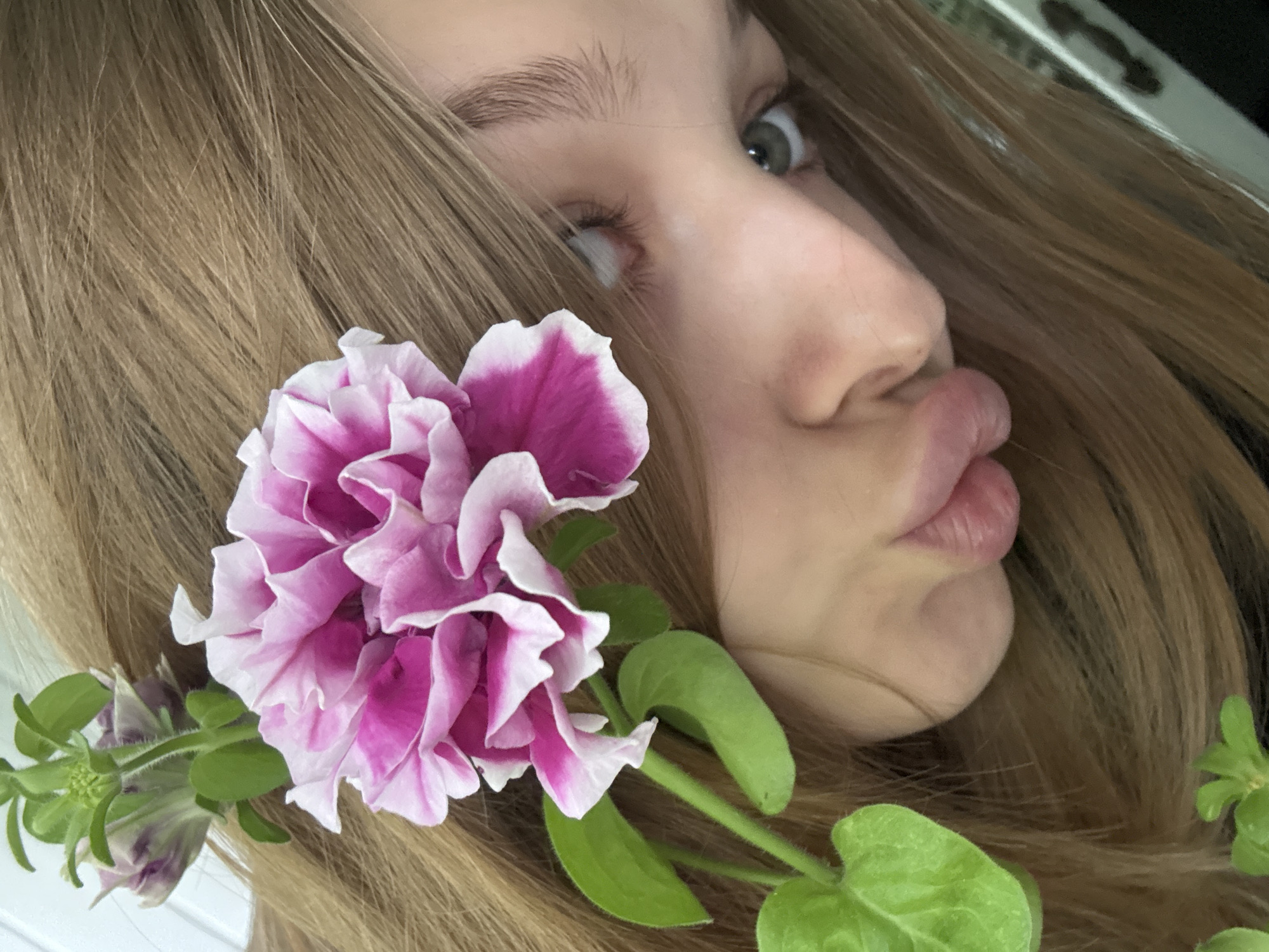 A close-up, angled portrait shot of a young person with long, light brown hair, identified as being in Chaikovsky, Russia. The subject's face is partially visible, showing one light-colored eye looking upwards and to the right, a visible eyebrow, and the nose. The lips are pursed in a distinct "pouting" or "kissing" expression. A large, double-petaled flower, pink with white edges, rests prominently on the subject's hair, slightly obscuring the other eye. A green stem and several bright green leaves extend from the main flower, with another smaller, light purple flower bud visible further back. The subject's hair forms the primary background, with a blurry, indistinct light area in the upper left and a dark, patterned object partially visible on the far right.