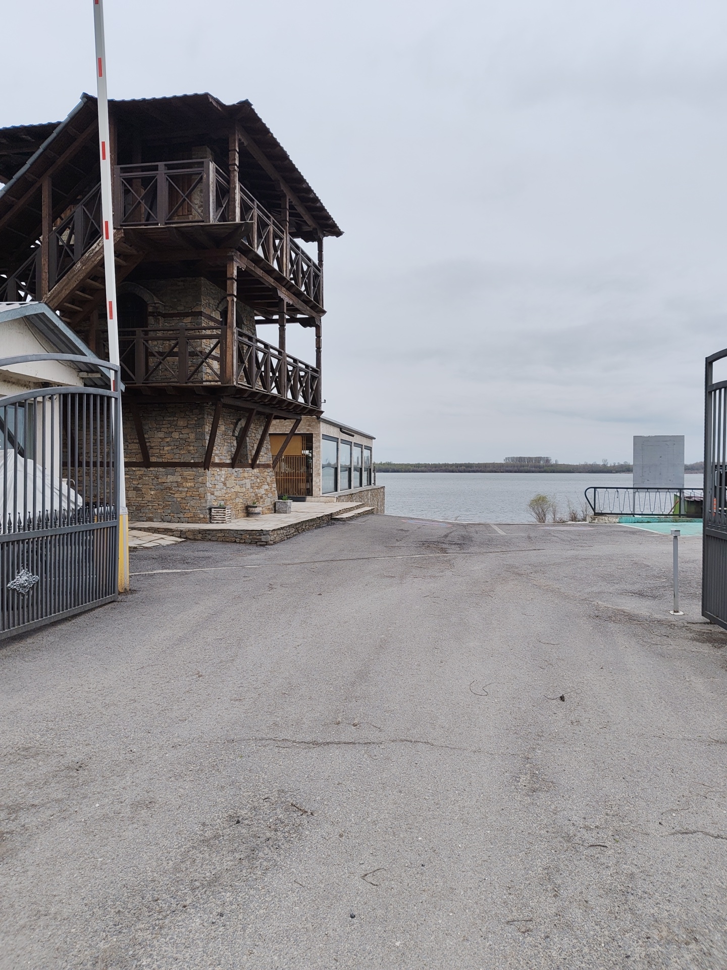 The image shows a building located in Kozloduy, Bulgaria, near the Danube River. The building has a three-story wooden tower section and a stone base. It is entered through an open gate with a partially visible white barrier. To the right of the building, a paved area leads towards the river bank, which is lined with some vegetation. Across the river, a line of trees is visible on the opposite shore. The sky is overcast.