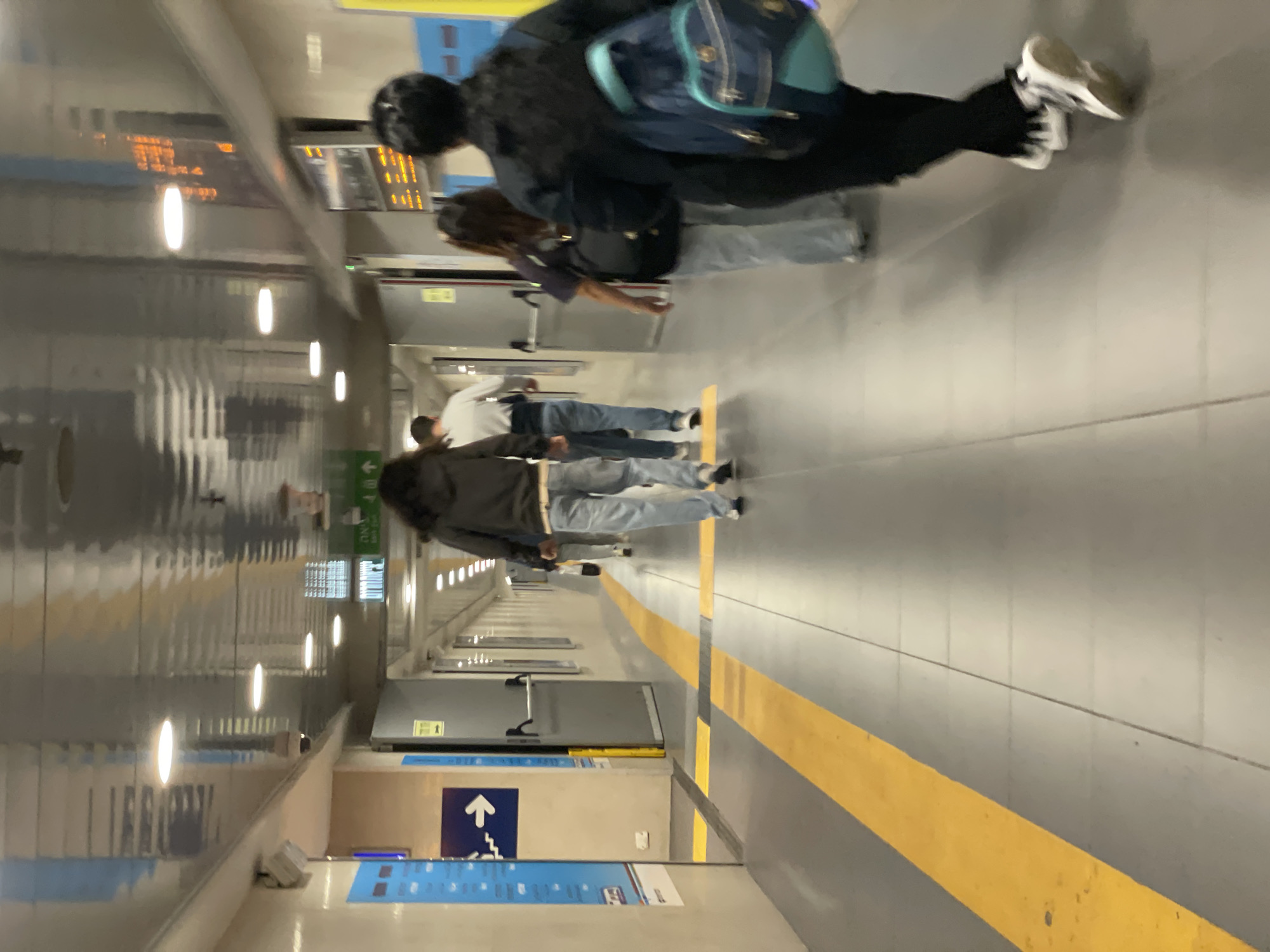 People are walking along a corridor in what appears to be a train station in Herzliya, Israel. The corridor has gray walls, a yellow safety line, and informational signs. There are other people further down the corridor.