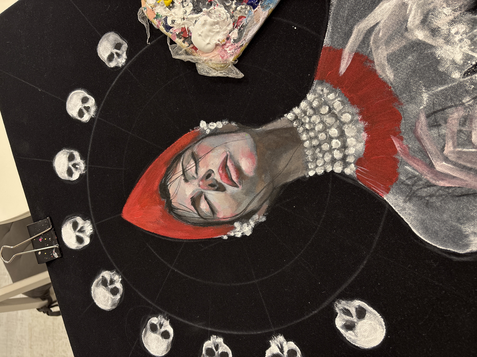 The image displays an in-progress painting rendered on a black fabric surface, likely a canvas or textile. The central subject is a stylized depiction of a person's face, oriented horizontally, with their head tilted slightly. The face, possibly a woman's, features pale skin, closed eyes with long lashes, and partially open red lips, conveying a serene or somber expression. She wears a prominent red hood or head covering that frames her face, and an elaborate, textured collar or necklace composed of numerous white or silver dots around her neck. Her garment, visible on the right, is painted in shades of grey and white with distinct brushstrokes.
Arranged in a circular pattern around the central figure are multiple small, white painted skulls, each rendered with simple detail. Thin white lines are faintly visible, radiating outwards from the center, indicating a guiding structure for the composition.
In the upper right portion of the image, an artist's palette is present, covered in plastic wrap and holding various paint colors including white, yellow, red, blue, green, and pink. A fresh dollop of white paint sits prominently on the palette. A black binder clip, speckled with paint, is visible on the left edge of the fabric, securing it. The overall artwork suggests a gothic, mystical, or Memento Mori theme. No visible text is present in the image.