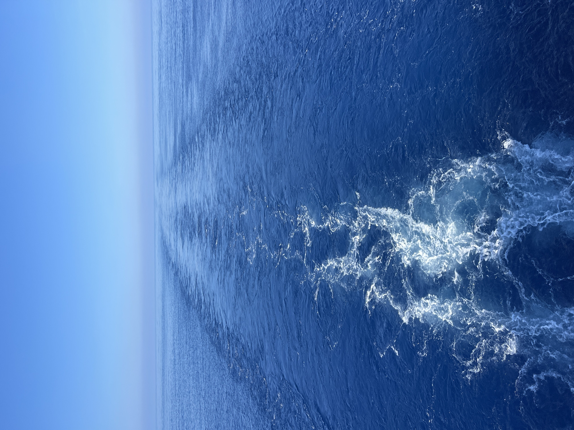 The image shows a wide expanse of deep blue ocean under a clear, pale blue sky. A ship's wake is visible, creating a foamy, white trail that recedes into the distance. The wake is widest in the foreground and narrows as it stretches towards the horizon, where the ocean and sky merge. The water is calm except for the turbulence of the wake, and there are no landmasses or other objects visible in the scene. The overall impression is one of vastness and open water.