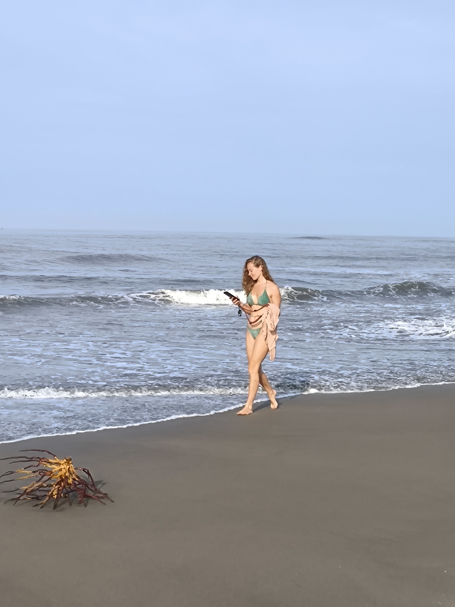 A woman with long curly hair, wearing a green bikini and a tan wrap, walks barefoot on a sandy beach alongside the ocean. She is looking at a small, dark object held in her hands, possibly a phone. The waves are visible on her left, and a cluster of seaweed lies near the water's edge. The sky is pale blue. The photo depicts a scene in Tibubeneng, Indonesia.
