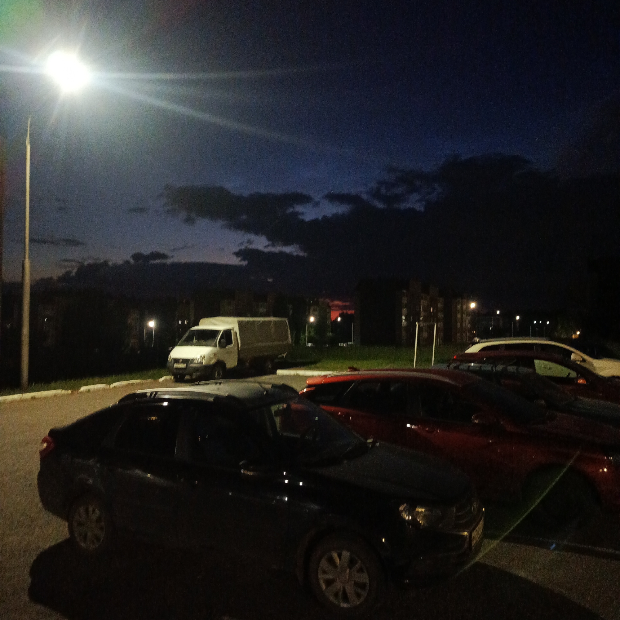 The image depicts a nighttime scene in a city, likely in Марковский, Russia, focusing on a street or parking area illuminated by a bright street lamp. Several vehicles are parked, including a white van with a covered cargo bed, a black sedan in the foreground, and red cars in the background. The sky is dark with visible clouds and a faint glow in the distance, suggesting dusk or dawn. The environment appears to be residential, with apartment buildings visible in the background. The lighting conditions create strong shadows and highlights, with the street lamp casting a glare across the scene. There are no people visible in the image.