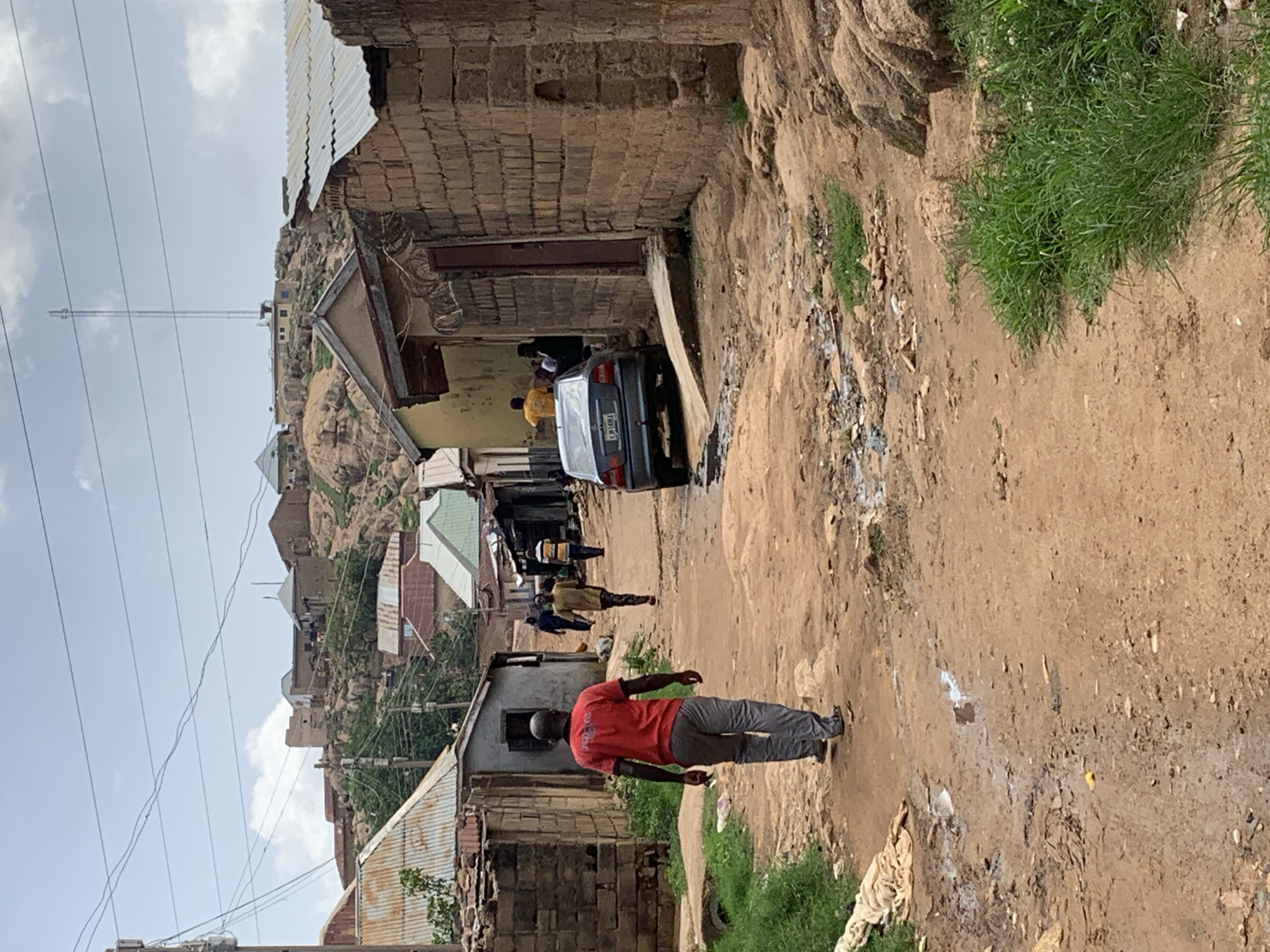 This image captures a street scene in Jos, Nigeria, characterized by an unpaved, dusty path winding through a residential area. The setting features a mix of informal and built structures, with a prominent rocky landscape in the background.
In the foreground and midground, several individuals are present. On the right side of the path, a man wearing a red t-shirt and grey trousers walks away from the viewer. Further down the path, a dark grey sedan car is parked on the left side, with at least two people standing near it; one is visible in a yellow shirt by the driver's side. Other figures can be seen walking further down the road, blending into the background.
The buildings lining the path and scattered across the rocky hills are constructed primarily from stone or brick, many featuring corrugated iron roofs. The terrain is rugged, with exposed rock formations and patches of green grass interspersed with bare earth and some debris. Power lines crisscross the sky, adding to the urban landscape. A tall communication tower or mast is visible on a distant hill, overseeing the area.
The time of day appears to be daytime, with visible clouds in a bright sky, suggesting either morning or afternoon. The overall impression is that of a bustling, lived-in community within a rugged natural environment.
A notable detail is the license plate of the parked car, which clearly displays "ABUJA" and beneath it "FCT".