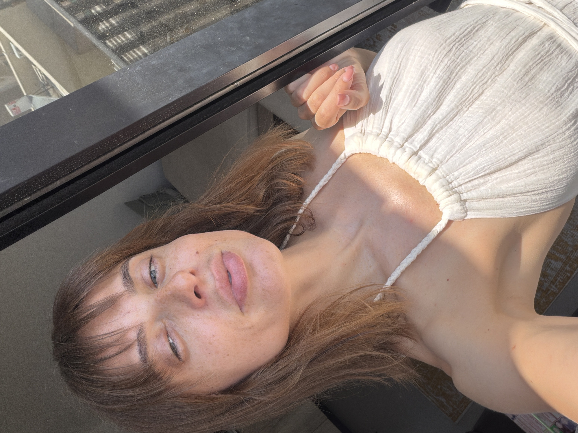 In a close-up selfie taken outdoors, a woman with light brown hair and bangs is visible from the chest up. She is wearing a white, textured dress with a halter neckline. The woman is looking at the camera with her left eye squinted and her lips pursed. Her skin is fair with visible freckles. Sunlight illuminates her face and upper body. She is standing near a dark-colored window frame. In the background, through the window, a building with balconies and railings is discernible, suggesting an urban environment. The scene appears to be a residential area in Tel Aviv, Israel, likely on a balcony or near a window.