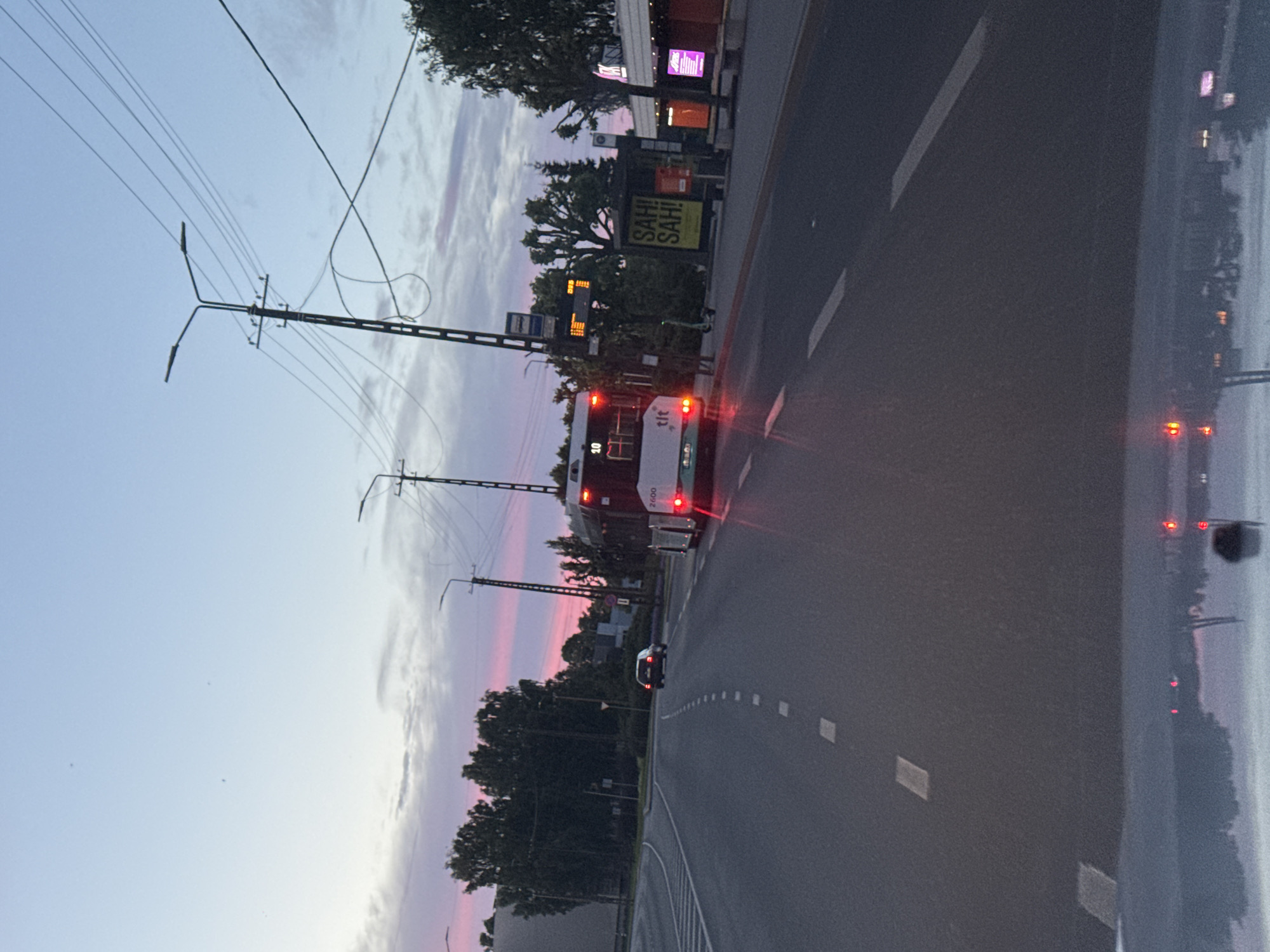 The image captures a street scene in Tallinn, Estonia, during twilight. A white bus with red taillights is visible on the right side of the road, appearing to be in motion. The bus has the number "10" displayed. To the left of the bus, a dark-colored car is traveling ahead on the road. The road is paved and marked with dashed white lines. Streetlights with multiple bulbs are positioned along the road, and overhead power lines are also present. In the background, trees line the side of the road, and buildings are visible, including one with illuminated signs that read "SAH! SAH! SAH!". The sky displays a gradient of soft pinks and blues, characteristic of sunset or sunrise. The scene is captured from the perspective of someone inside a vehicle, as suggested by the reflection of lights and the hood of a car at the bottom of the image.