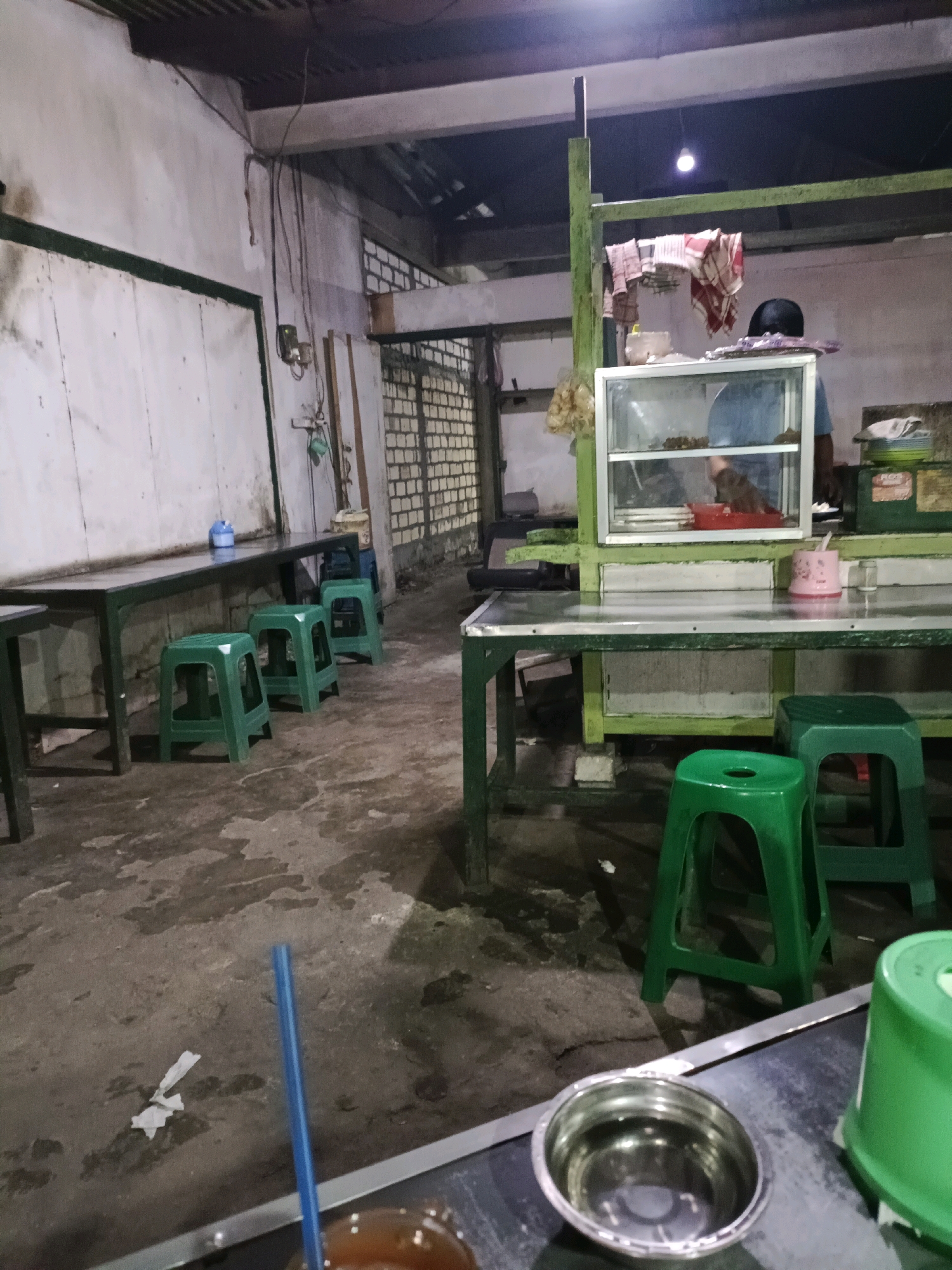 The image depicts an outdoor food stall or small eatery in Sukomulyo, Indonesia. The scene is dimly lit, suggesting it is either early morning or evening. In the foreground, a metal counter holds a bowl of water and a blue straw, with a green plastic container partially visible to the right. The floor is concrete and appears somewhat damp, with some debris scattered around.
The stall itself has a green metal frame supporting a glass display case filled with food items, with a person in a blue shirt and black cap partially visible behind it, attending to the food. Red and white checkered cloths are hanging from the frame above the display. Various green plastic stools are placed around tables with metal surfaces.
The background shows a white wall with a green border, and beyond that, a wall constructed from light-colored bricks. There are electrical wires and some lighting fixtures visible, indicating the operational nature of the establishment. The overall impression is of a functional, no-frills dining area, likely catering to a local clientele.
