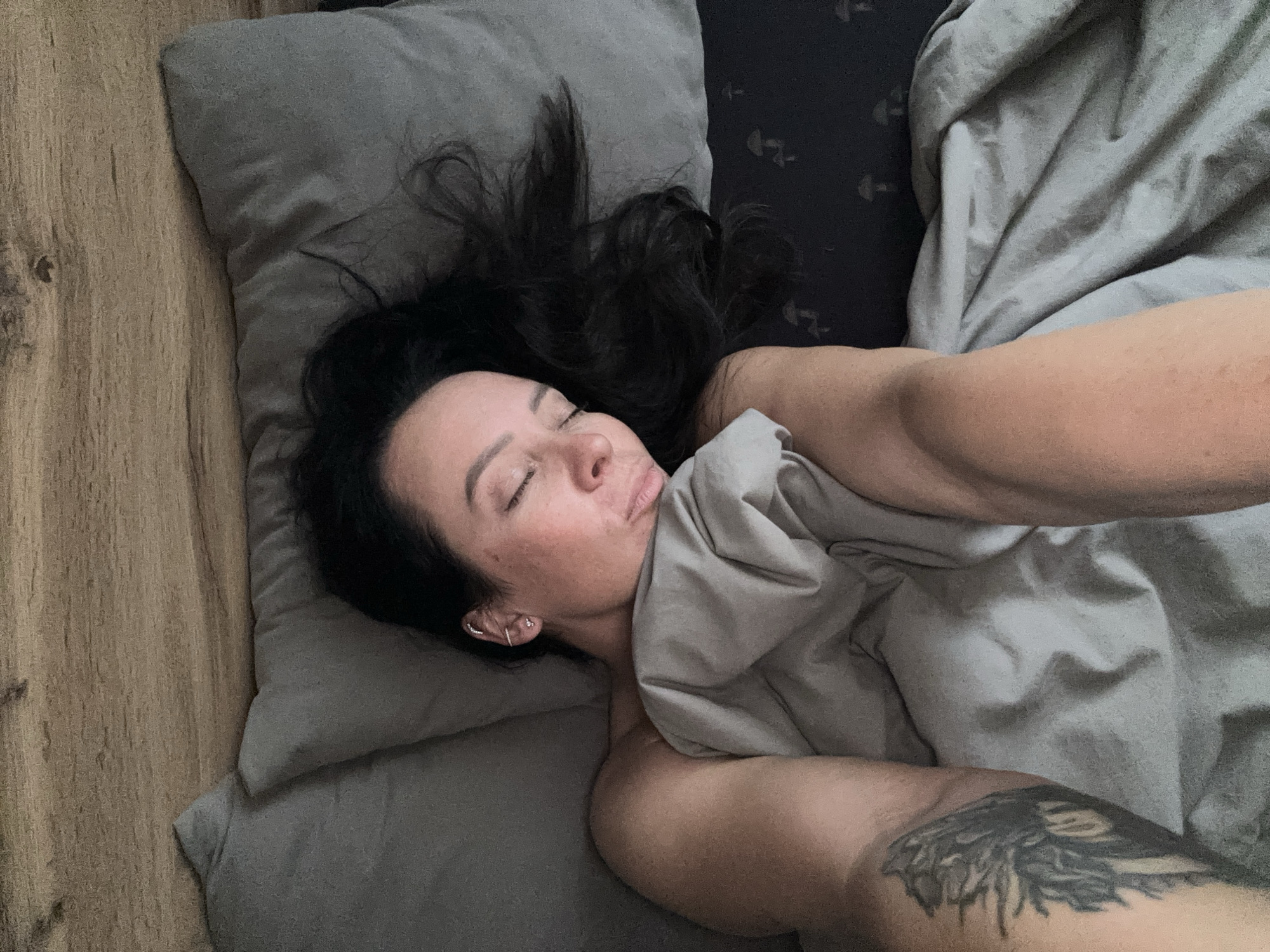 A woman with long dark hair is lying on a bed with gray bedding and a pillow. Her eyes are closed, and she appears to be sleeping. She has light skin, and a tattoo is visible on her arm. The bed is on a wooden floor. There is no visual indication of the location being Chaikovsky, Russia.