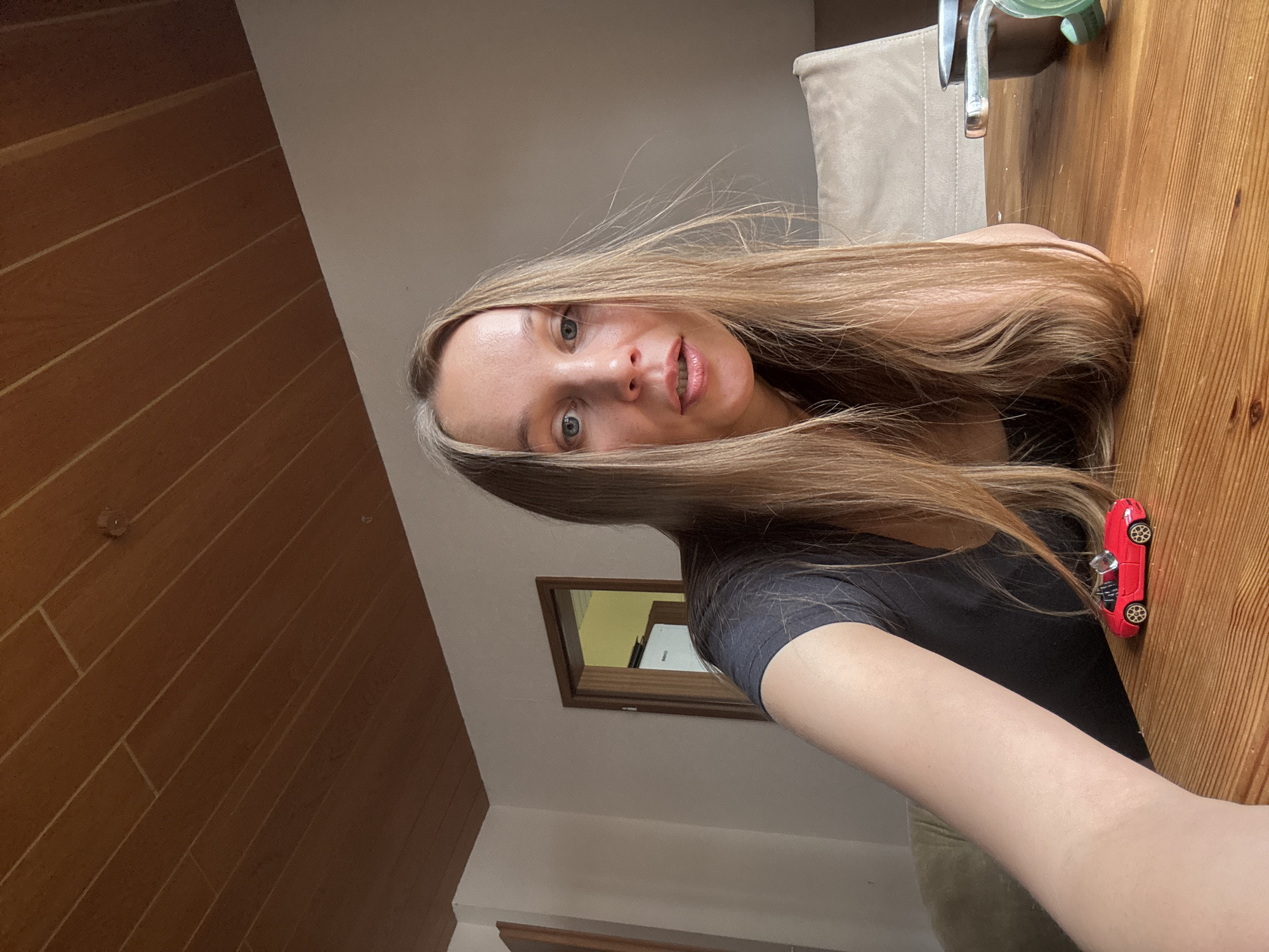 A young Caucasian woman with long blonde hair and blue eyes is captured in a selfie taken indoors at a wooden table. She is wearing a dark t-shirt and is positioned in the foreground of the image, looking directly at the camera. Her lips are slightly parted, and her facial expression appears natural.
On the table in front of her, there is a small, bright red toy convertible car. To the right of the woman, partially out of frame, a tan-colored chair back is visible, suggesting she is seated at a dining or kitchen table.
The background consists of a plain, light-colored wall and a wooden paneled ceiling. A doorway is visible on the left side of the background, leading into another room with a white appliance and a yellow-lit area. The scene is well-lit, with natural light appearing to come from the right side of the frame. The overall environment suggests a domestic setting, possibly a home in Überherrn, Germany, though no specific landmarks or city indicators are present in the image itself. The focus is on the woman and the toy car on the table.