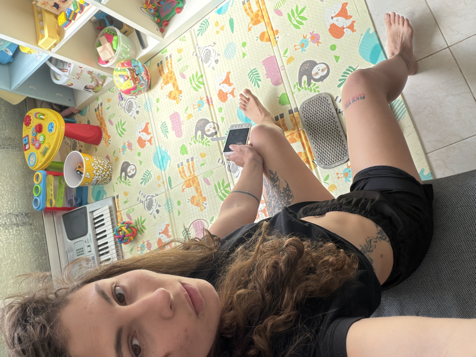 A young adult female is depicted lying down indoors, taking a selfie with a smartphone. She has long, curly brown hair, dark eyes, and a neutral, relaxed expression. She is dressed in a black t-shirt and black shorts. Visible tattoos include floral designs on her left arm and abdomen, and a prominent tattoo on her left thigh that reads "FUCK YOU" in stylized blue/purple block letters.
She is resting on a gray sofa or cushion, with her legs extending onto a colorful foam playmat that covers the floor. The playmat features a light yellow background adorned with cartoon illustrations of various animals such as giraffes, zebras, foxes, sloths, birds, and elephants, interspersed with green leaves.
The surrounding environment indicates a child-friendly space, likely a home in Kiryat Bialik, Israel. A white shelving unit in the background holds an assortment of toys, including building blocks, buckets, and a colorful ball. On the floor near her, various other children's items are present, such as a yellow activity center, a red cone-shaped toy, a cup with a toy car, a small electronic keyboard (Casio CTK-50H), and a colorful rainbow ball. To the left of her left leg, a gray Sadhu board (bed of nails) is placed on the playmat. The lighting suggests it is daytime.