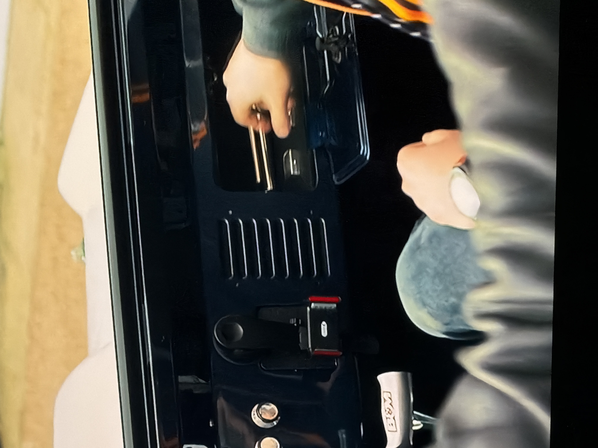 The image captures a close-up view of a person interacting with the interior of what appears to be a vehicle, possibly the tailgate or a storage compartment of a truck or SUV, located in Sofiivska Borshchahivka, Ukraine. The scene is dominated by dark blue or black surfaces of the vehicle's interior.
Two hands of a person are visible. The left hand, wearing a dark green sleeve, is reaching into an open compartment, holding several long, thin, metallic objects, possibly keys or small tools. The right hand, also with a dark green sleeve, is holding a white or silver can, which could be an aerosol spray or similar container.
Notable details of the vehicle's interior include a section with vertical slatted vents, a black latch mechanism with red accents, and two round, silver-colored buttons or fasteners below the latch. To the lower right, a silver-colored handle or knob is visible, clearly etched with the text "B&M."
The background beyond the vehicle shows a light-colored surface, possibly a wall or structure, and a strip of a light brown, wooden texture, suggesting the vehicle might be parked outdoors or in a garage. The lighting is focused on the immediate area of activity, creating a somewhat dark and intimate setting for the task being performed. No strong emotions are discernible from the hands.