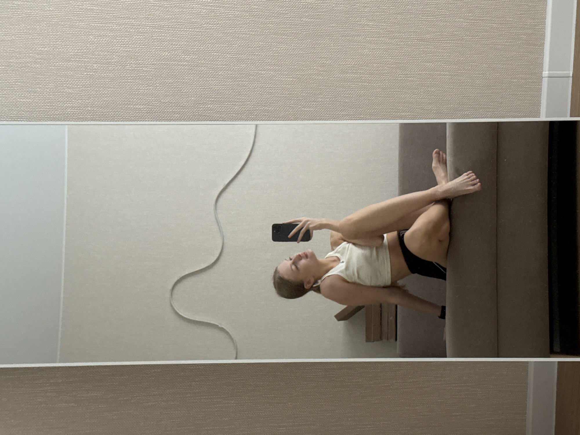 A young woman is captured in a full-length mirror selfie, posing on a brown couch. She is wearing a white crop top and black shorts. Her legs are crossed, and she is holding her phone up to take the picture. The background of the room appears to be a neutral color, with a decorative wave pattern on the wall behind her. The image is a straightforward photograph without any specific identifying features that would place it in Perm, Russia.