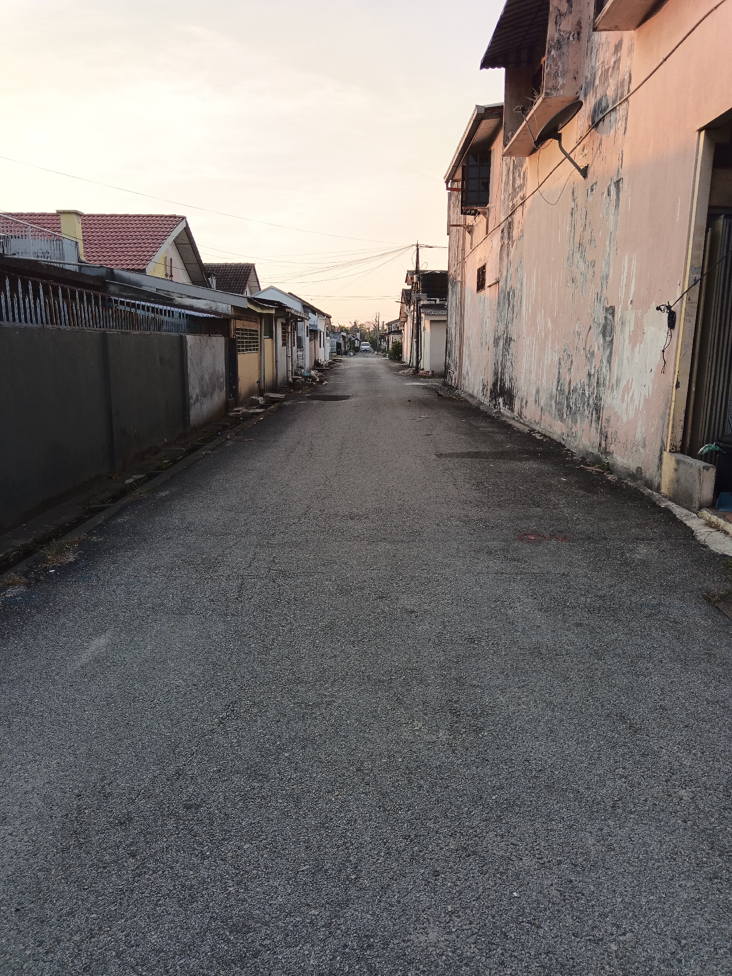 The image depicts a street scene in Klang City, Malaysia, likely during the late afternoon or early evening, given the soft, warm light. The perspective is from the middle of an asphalt road, looking down a moderately wide street lined with residential buildings on both sides.
On the left side of the street, there is a row of houses with tiled roofs, some appearing to be single-story dwellings with fences and walls in front. The buildings on this side are varied in color and design, suggesting a mix of older and newer constructions. Power lines are visible crisscrossing above the street and buildings.
The right side of the street features a prominent building with a light pink, weathered facade. The wall shows signs of age and disrepair, with peeling paint and stains. An exterior satellite dish is mounted on this wall, along with electrical conduits and wiring. Further down this side, other buildings with corrugated metal roofs and various architectural styles are visible. A metal gate and some debris, including a green plastic trash bag, are present near the foreground on the right.
The road surface is asphalt, appearing somewhat worn in places. There are no people or vehicles immediately visible in the foreground, though a white car can be seen in the distance at the end of the street. The overall impression is one of a quiet, established residential neighborhood in a Malaysian city.