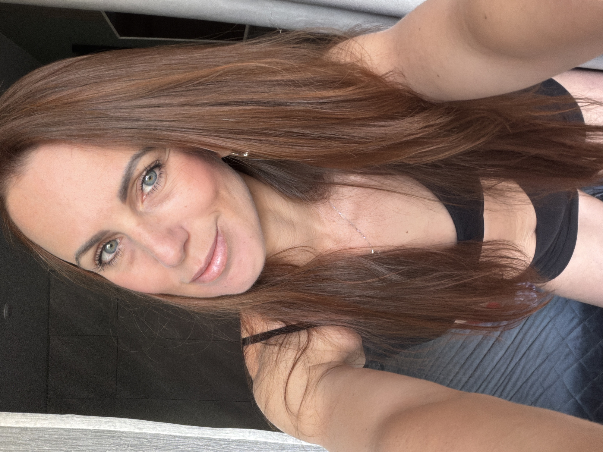 The image is a close-up, selfie-style photograph of a woman with long, reddish-brown hair. She has blue eyes and is wearing a black bra and black underwear. She is indoors, possibly in a bedroom, with a quilted dark grey surface visible in the background, suggestive of a bed. Curtains or drapes are visible on the left and right sides of the image. The lighting suggests it is daytime, possibly near a window. There is no indication of the location being in Birobidzhan, Russia.