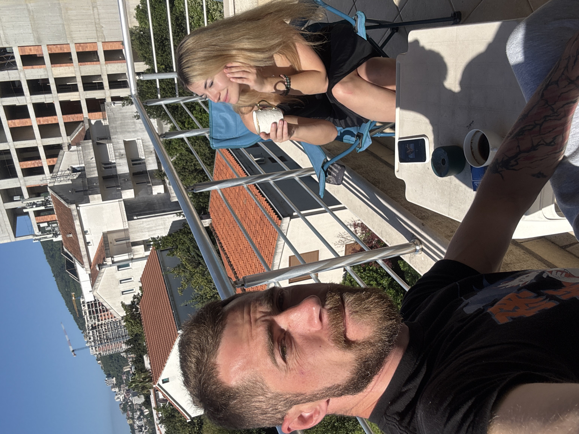 A man takes a selfie on a balcony in Budva, Montenegro. A woman sits behind him, holding a white cup. A small table holds a phone, a green speaker, and a coffee cup. In the background are buildings, including a new construction site with a crane. The weather is sunny and clear.
