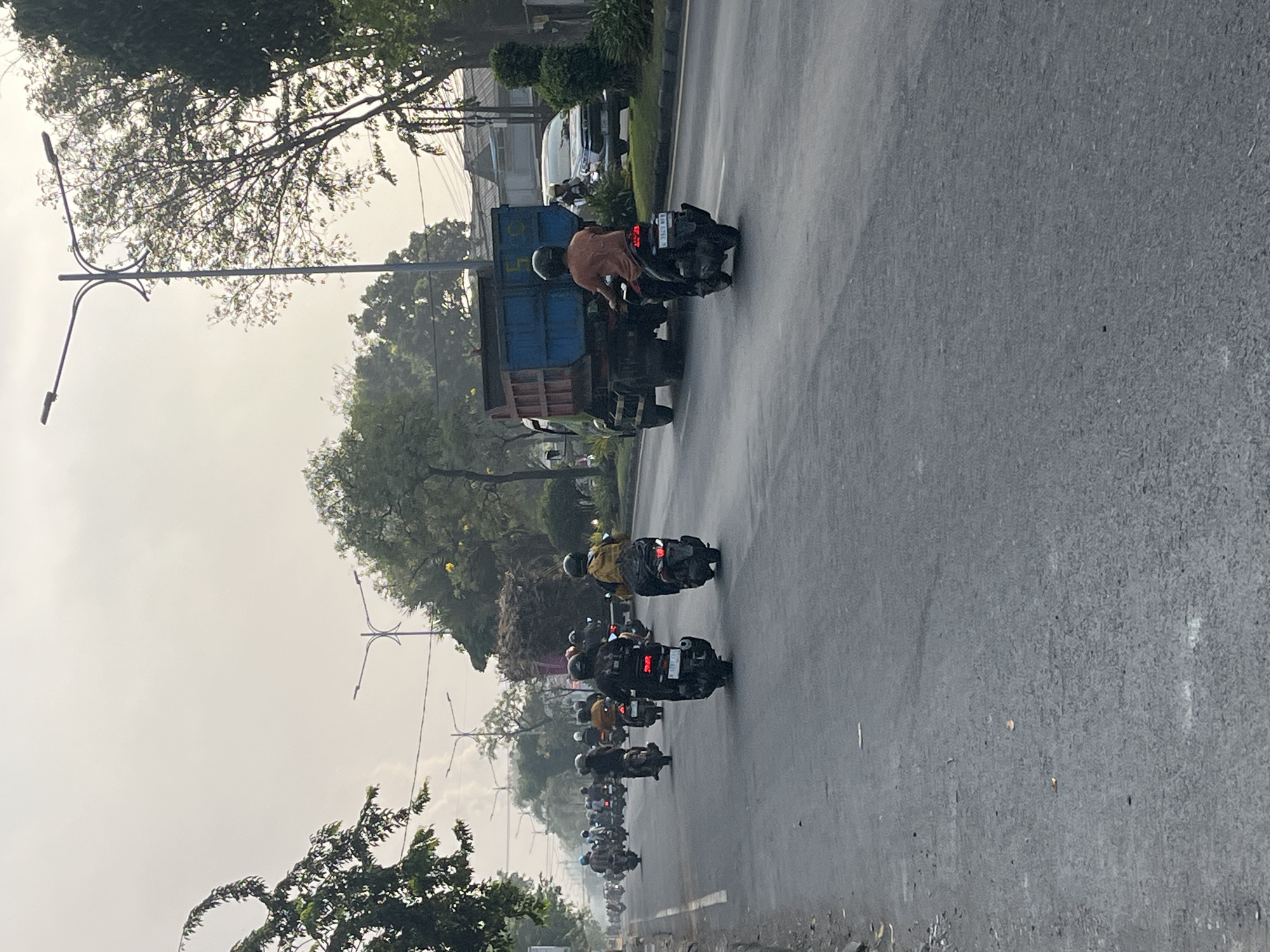 The image depicts a street scene in Gelam, Indonesia, likely during daylight hours. A traffic jam of motorcycles and a truck is visible on a paved road. The sky is overcast, and trees line the sides of the road. Several motorcycles are in motion, with riders wearing helmets. One rider in the foreground is on a black motorcycle, wearing an orange shirt and a helmet. Behind them, a blue and green truck carries a load of hay or similar material. A white car is parked on the right side of the road. Streetlights are visible in the background. The overall impression is that of a busy urban or semi-urban environment.