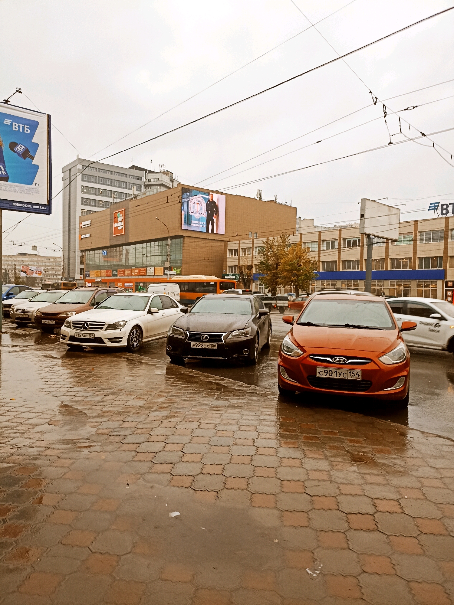The image depicts an outdoor scene in a city, likely on a cloudy or overcast day, with wet ground suggesting recent rain. Several cars are parked and visible on a paved surface. In the foreground, an orange Hyundai is prominently featured. Behind it, a white Mercedes-Benz sedan is parked, and further back, a dark-colored sedan, possibly a Lexus, is visible. The background shows urban buildings, including a multi-story structure with a large digital billboard displaying an image of a man in a uniform. Power lines are strung overhead. The paving stones on the ground are wet and reflective.