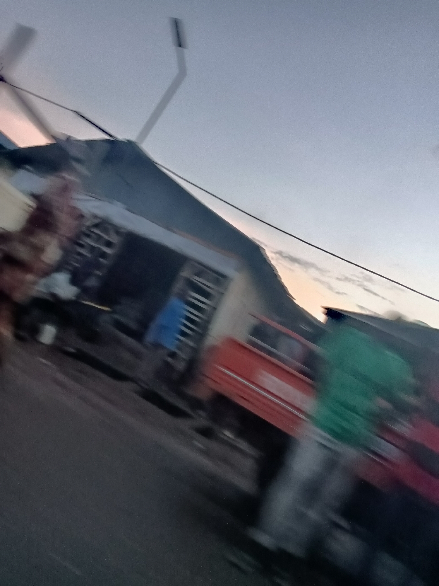 This is a motion-blurred, eye-level, exterior shot taken at dusk in Jos, Nigeria. The scene appears to be a street with buildings and a few vehicles. In the foreground, a red truck is partially visible. To the left of the truck, a person wearing a green shirt is standing. Behind the truck, a building with an open front is visible, and another person wearing a blue garment is inside. The sky is clear with faint clouds, and a power line stretches across the frame. The overall impression is of a busy street scene captured during a low-light condition with significant motion blur.