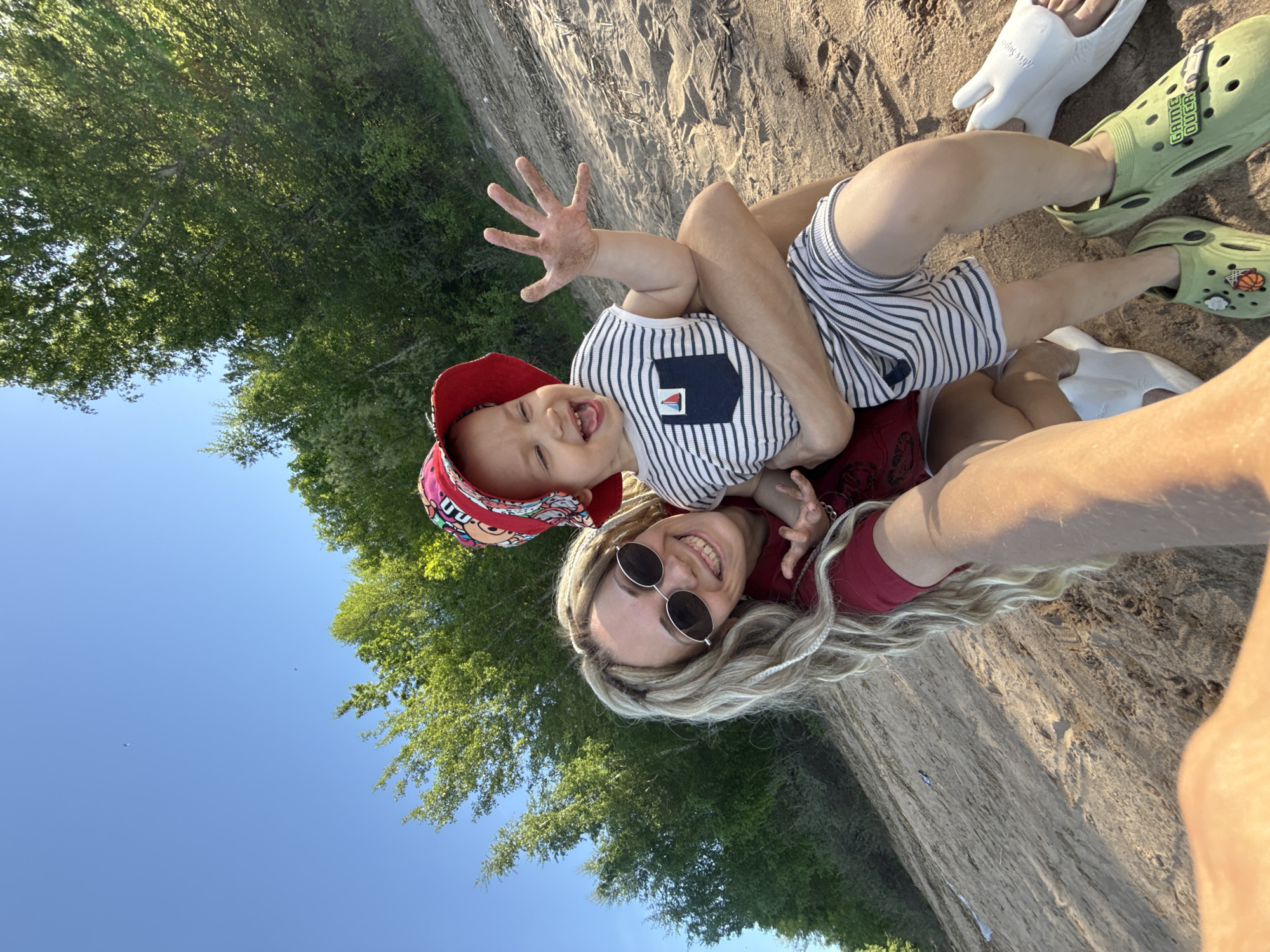 A photograph depicts a woman and a young child outdoors, possibly in Шебунино, Russia. The woman is lying on the ground, wearing a maroon top, sunglasses, and has blonde hair. She is holding the child. The child is wearing a striped shirt, striped shorts, and a red hat. They are smiling and appear to be interacting playfully. The child's hand is raised, covered in sand. The background includes a sandy area and trees. The sky is visible in the corner. The photograph appears to be taken from an angle that gives a tilted perspective of the scene. Footwear is visible: the woman is wearing white sandals and the child is wearing green Crocs.
