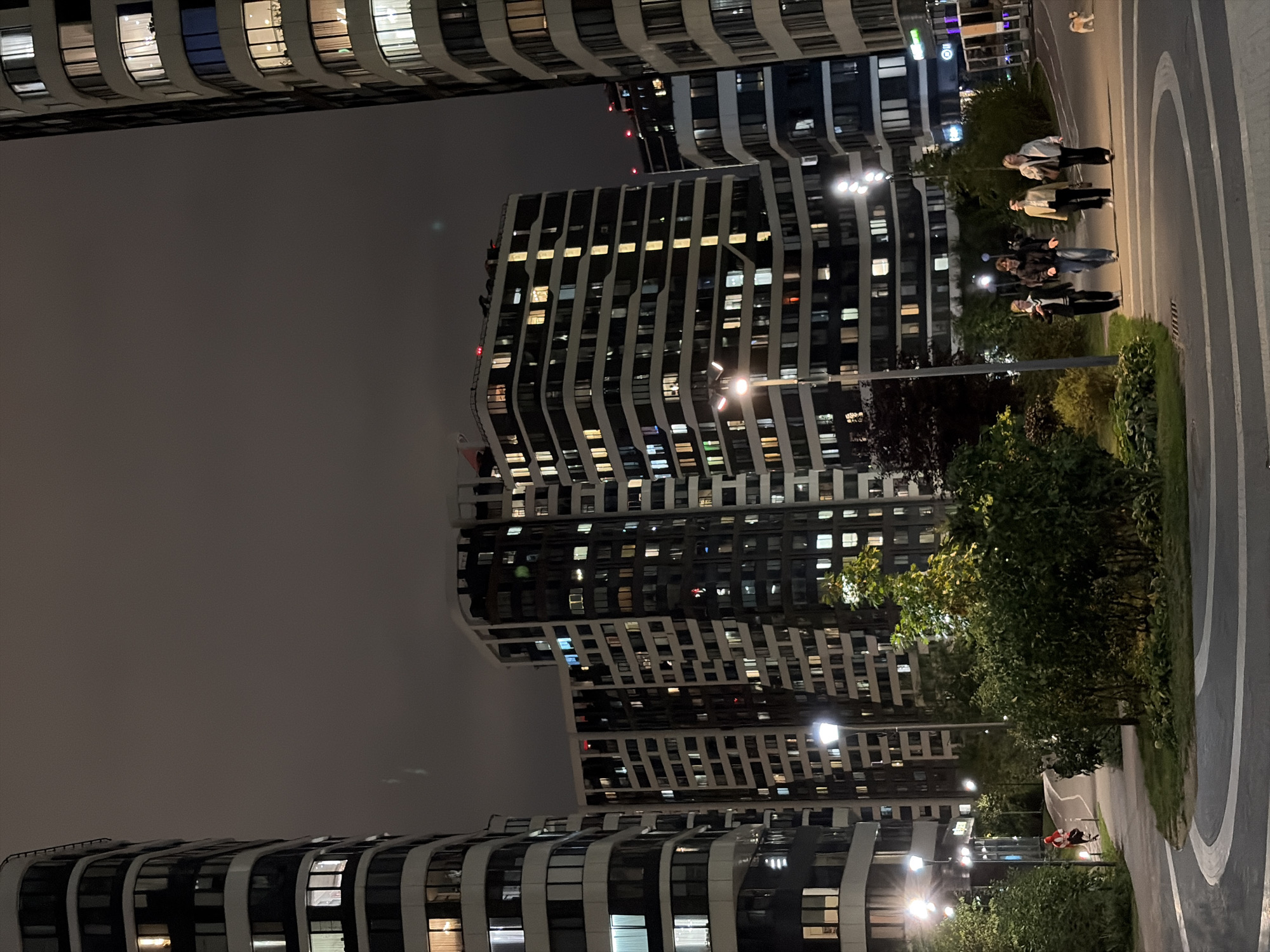 In a residential complex in Moscow, Russia, at night, several modern residential buildings stand tall, their windows illuminated with warm light. A pathway curves through a landscaped area with trees and bushes, separating the buildings from the walkway. Four young people are walking on the pathway, their figures silhouetted against the light. To the left, a person is running on a different path. A small dog is visible in the distance on the right. The sky is dark and cloudy, typical of an overcast evening in Moscow. The scene captures a moment of urban life at night, with people engaging in recreational activities amidst the illuminated architecture.