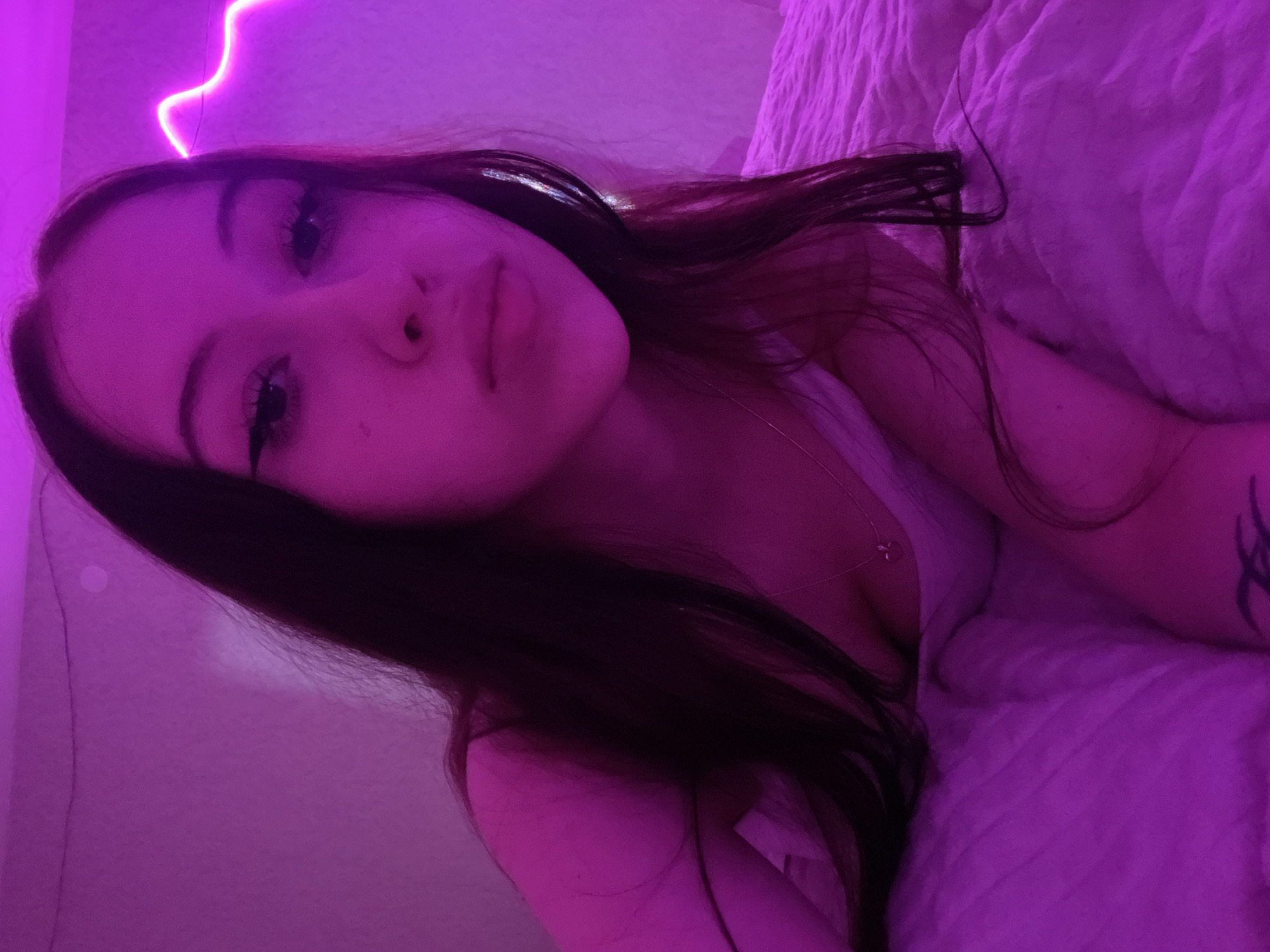 A young woman with long dark hair is lying on a pink blanket under purple lighting. She is wearing a white top and a necklace. A neon pink light strip is visible in the background. The scene appears to be indoors, possibly a bedroom.