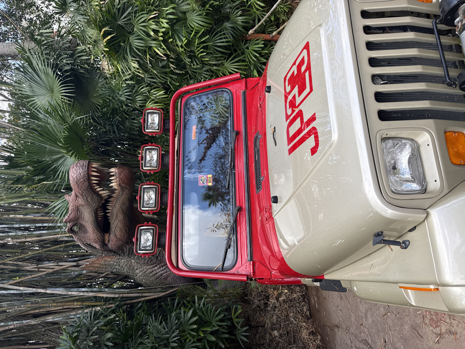 The scene depicts a static outdoor display at Universal's Islands of Adventure in Orlando, Florida, United States. A beige and red Jeep Wrangler (YJ model) is positioned centrally, facing forward. The vehicle's hood is tan/beige with the red text "JP23" outlined in white. Its upper body and roll bar are red, and a roof-mounted light bar features four rectangular auxiliary lights in red housings. A "Jurassic Park" logo sticker and the number "023" are visible on the upper windshield. Directly behind and above the Jeep, partially obscured by dense tropical vegetation including bamboo stalks and large palm fronds, is a life-sized animatronic or statue of a large theropod dinosaur, appearing dark brown/green. The dinosaur features prominent sharp teeth, visible green eyes, and its mouth is open. The ground visible to the left is reddish-brown earth. No individuals are present.