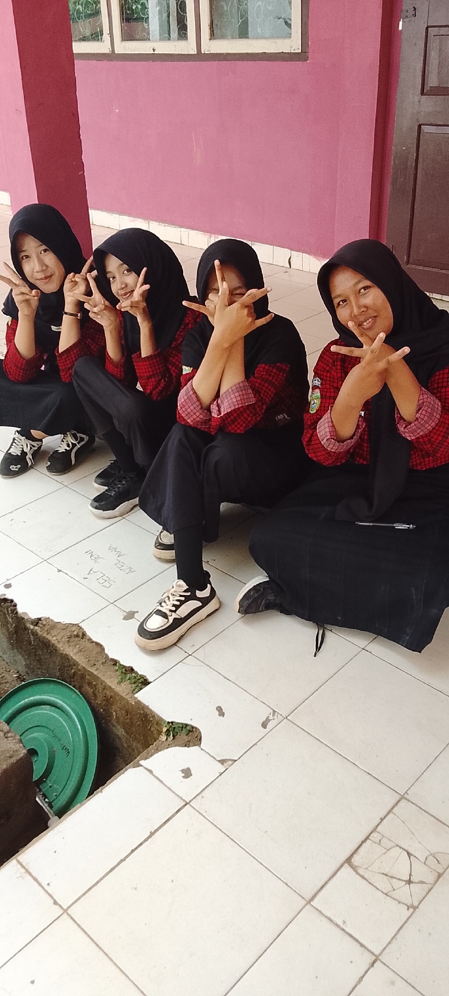 Four young Indonesian women, appearing to be in their late teens or early twenties, are sitting on a tiled floor in front of a pink wall. They are all wearing black hijabs and red and black plaid shirts. They are posing for a photo, with three of them holding up two fingers in a peace sign gesture. The woman on the far right is also making a peace sign gesture. The floor is made of white tiles, some of which are cracked or broken. There is a green circular object visible in a drainage ditch to the left of the frame. The background shows a pink wall with windows and a wooden door. The overall scene appears to be in an outdoor, possibly school or public, area.