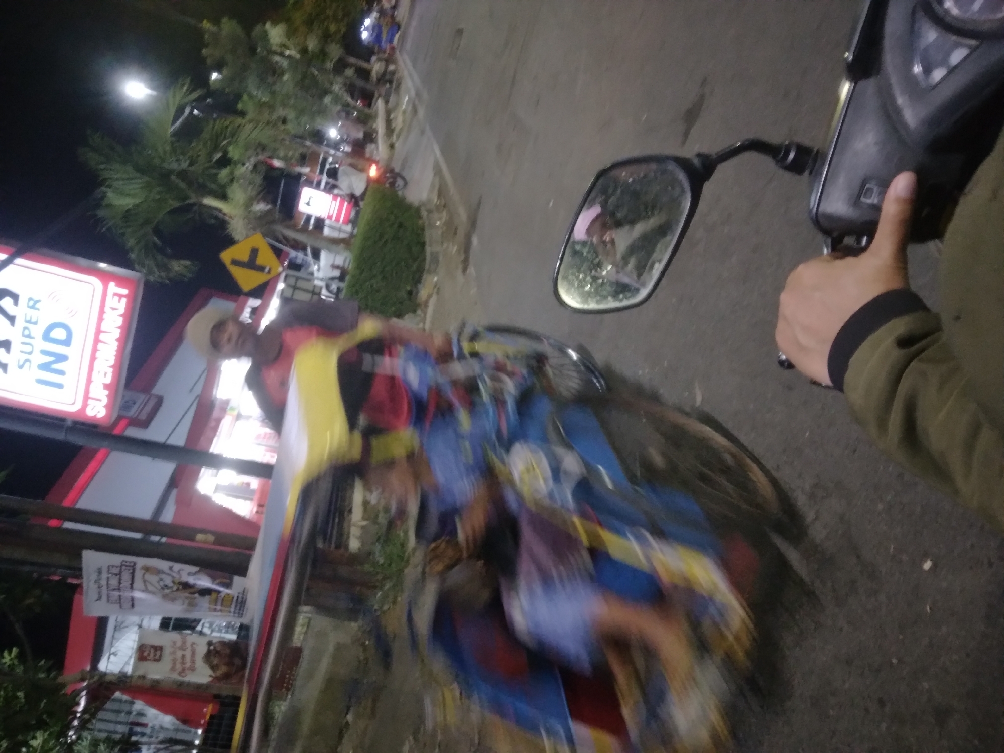 The image shows a scene at night in the city of Purwokerto, Indonesia. In the foreground, a person's hand is visible, holding the handlebar of a motorcycle. The motorcycle's mirror reflects a person wearing a pink headscarf. To the left, a blurry becak, a three-wheeled passenger vehicle, is in motion. The becak is carrying passengers, one of whom appears to be a child. In the background, a Super Indo supermarket is illuminated, with a sign displaying "SUPER INDO" in large letters. Other businesses and trees are also visible, along with streetlights and passing traffic. The overall impression is a busy street scene in an urban environment at night.