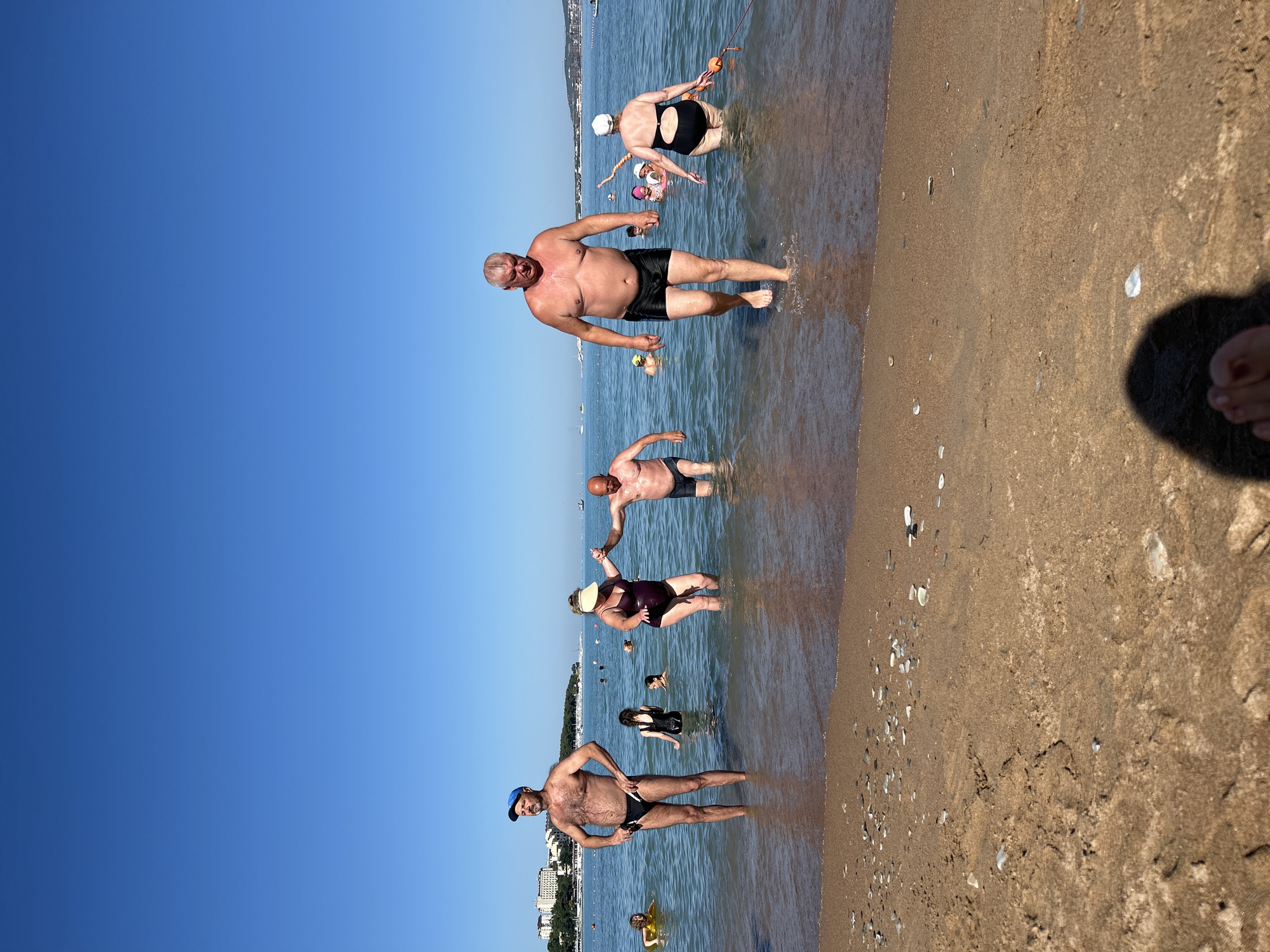 A group of people are standing in the shallow water at a beach in Gelendzhik, Russia, on a sunny day with a clear blue sky. In the foreground, the sandy beach has small pebbles and shells. The water is calm and clear.
From left to right, a man wearing a blue baseball cap and black swim briefs stands looking to his left. Next to him, a woman in a dark swimsuit is partially submerged in the water, holding hands with a man in dark swim trunks who is also in the water. Further to the right, another man, wearing black swim trunks, is walking out of the water towards the shore. Behind him, a woman in a black swimsuit with a white cap is standing in the water, holding a buoyant orange object. In the background, several other people are visible in the water, some swimming and some standing. In the distance, buildings and boats can be seen along the coastline, suggesting an urban or resort area. The overall scene depicts a casual day at the beach.