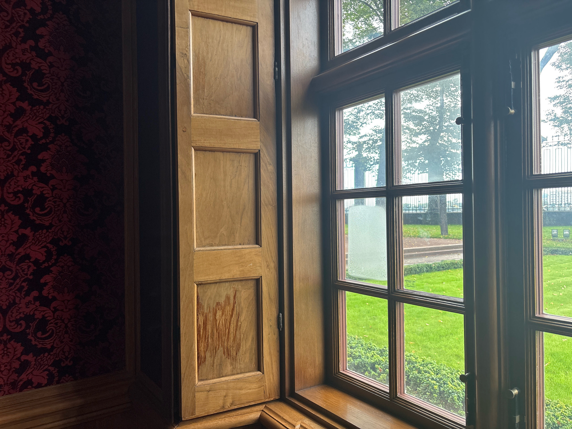 The image shows an interior view looking out of a large, multi-paned window. The window frame is made of dark wood, and the panes are divided into a grid pattern. Outside the window, a green lawn is visible, with trees and a path. The sky appears overcast. To the left of the window, a wooden shutter is partially open, revealing its natural wood grain. Behind the shutter, a wall is covered in dark red patterned wallpaper. The scene appears to be from a historic building, possibly a palace or museum, given the ornate wallpaper and wooden architectural details. This could be a room in a location like the Hermitage Museum or another grand estate in Saint Petersburg, Russia.