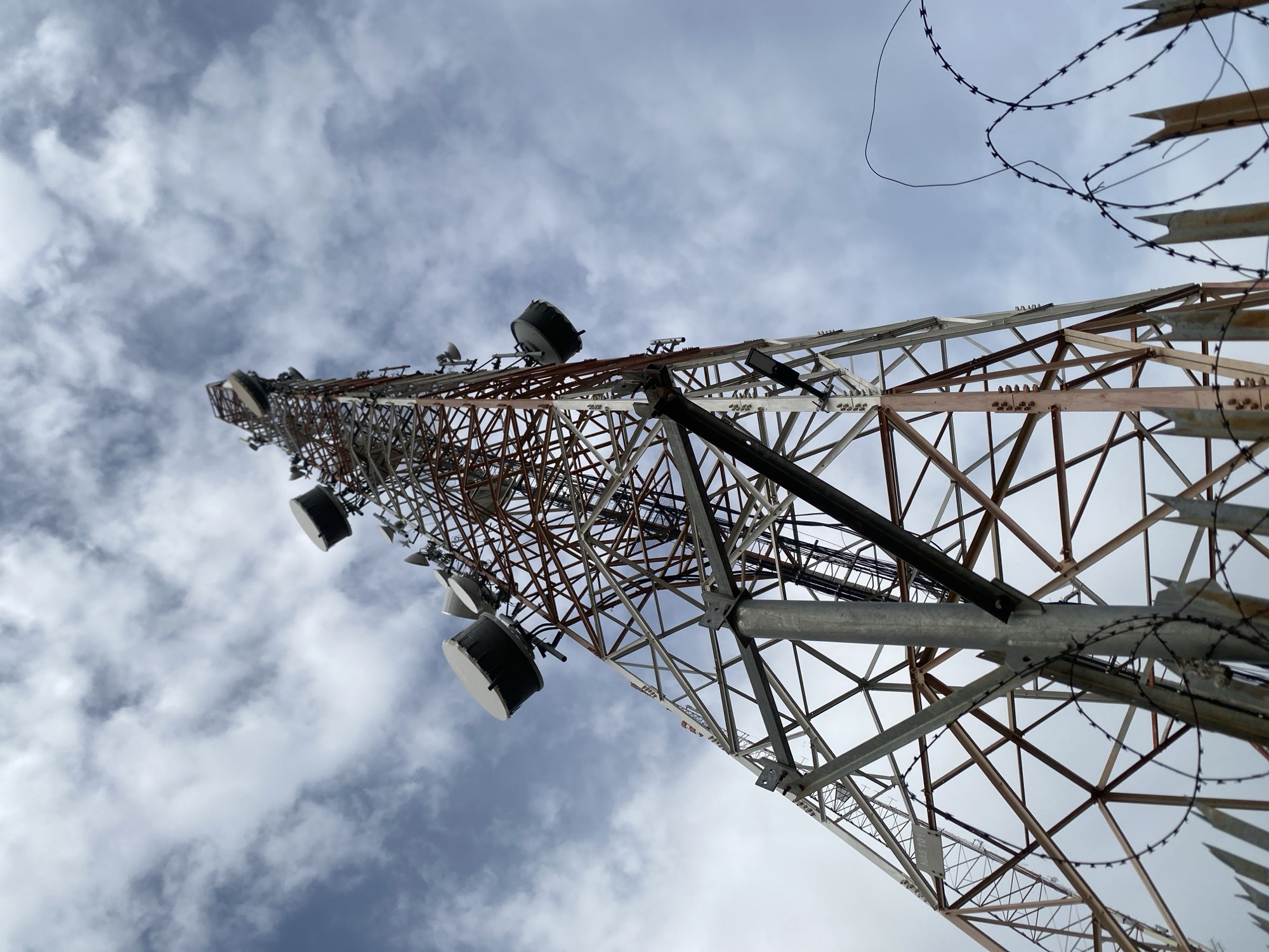 The media file shows a low-angle shot looking up at a cell tower against a cloudy sky. The tower is made of a lattice of metal beams, some painted red and others silver. Various communication equipment, including large cylindrical antennas, are mounted on the tower at different levels. Barbed wire is visible along the top of the tower, suggesting security measures. The sky is filled with white and gray clouds, indicating an overcast or partly cloudy day. The image appears to be taken during daylight hours. The scene is likely an outdoor location, possibly in a somewhat remote or industrial area, given the nature of a cell tower. No people are visible in the image. There is a small, white label attached to the tower with black text on it, but the text is too blurry to be read. The location is identified as Keffi, Nigeria.