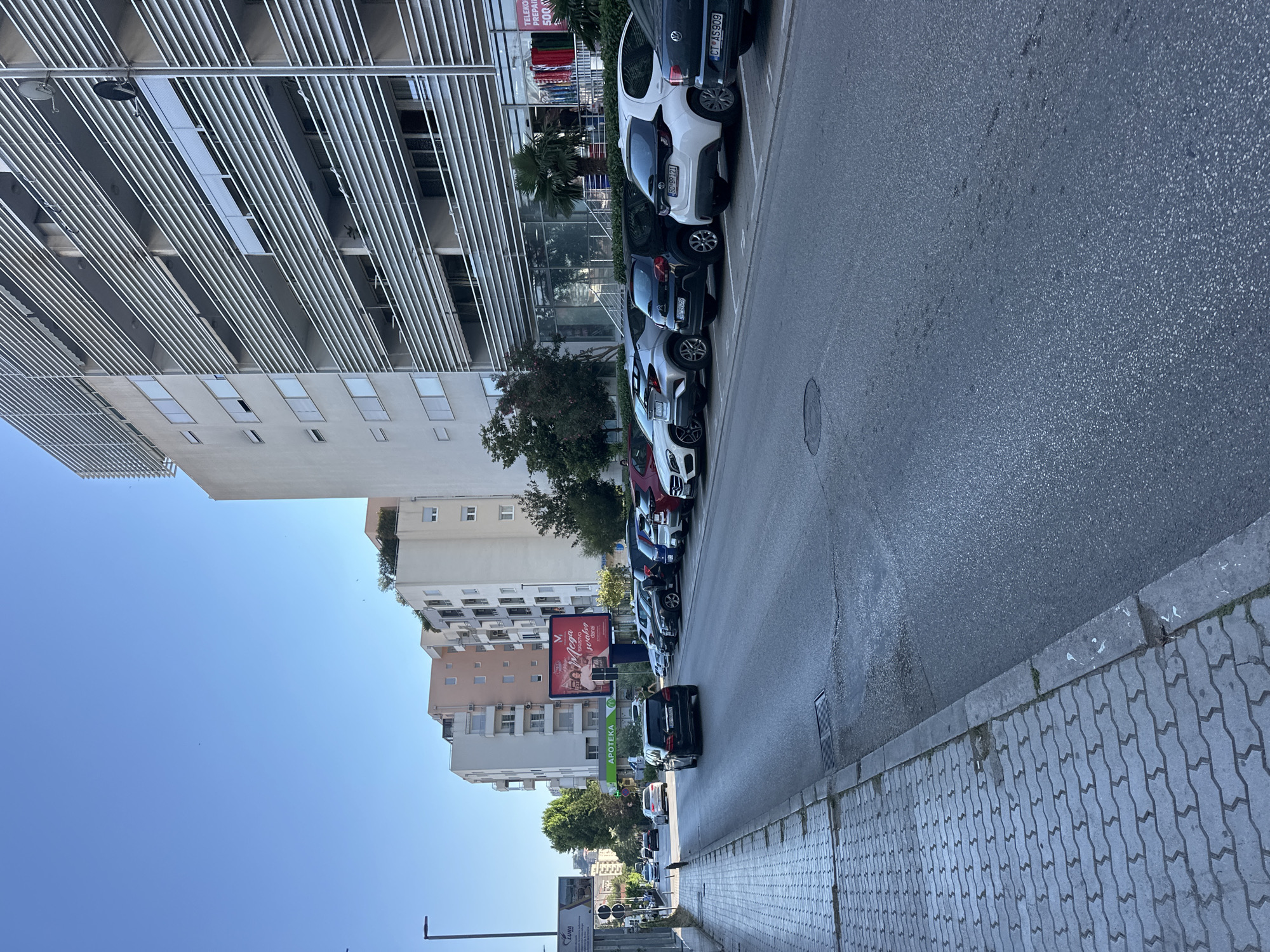 This is a ground-level, eye-level, exterior photograph taken on a sunny day, likely in the afternoon given the shadows. The scene depicts a street in Budva, Montenegro, with several cars parked along the right side of the road. The cars are a mix of makes and models, and some have visible license plates, such as "BD BR121", "CT AS909", and "50181059". The road itself is paved, and to the right of the road is a pedestrian sidewalk made of interlocking concrete pavers.
On the left side of the street, there are multiple buildings. The closest building appears to be a modern structure with a facade of horizontal silver louvers and recessed windows. Further down the street, there are several taller residential buildings with balconies. One building features a large billboard with text that appears to be in a Slavic language, including the words "Mega" and "APOTEKA", which means "pharmacy". There are also trees and palm trees visible, suggesting a Mediterranean climate. The sky is clear and blue. The overall atmosphere is one of a typical urban street scene in a coastal European town.