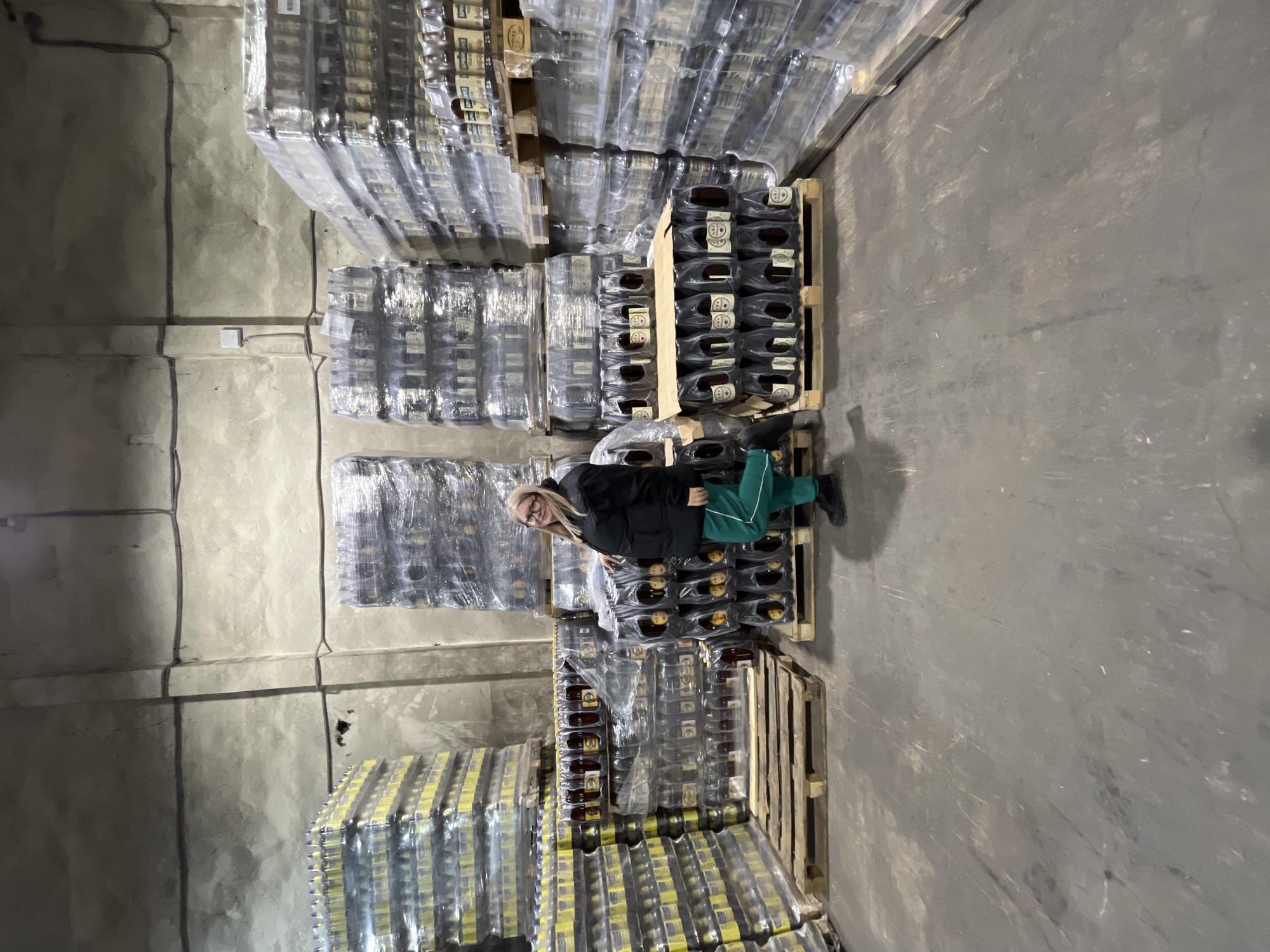An indoor photograph captures a female individual posing in a warehouse located in Izhevsk, Russia. The setting features light gray, unfinished concrete walls with visible conduit lines and a worn, dark gray concrete floor. The warehouse interior is extensively filled with wooden pallets stacked high with shrink-wrapped commercial beverages.
To the left, numerous pallets hold tall stacks of plastic bottles with yellow caps and yellow labels, suggesting a light-colored beverage. In the center and extending to the right, many pallets are laden with darker-colored bottled beverages, primarily in brown glass or plastic bottles, organized in multi-packs and secured with shrink wrap. Cyrillic text on some labels identifies these products as "Kvass."
A single female subject, with blonde hair and glasses, stands in the center-right of the frame, smiling directly at the camera. She is dressed in a black padded jacket, green track pants with a white stripe down the side, and black shoes. Her right arm rests upon a stack of the darker bottled beverages, and her left leg is casually bent with the foot slightly raised.