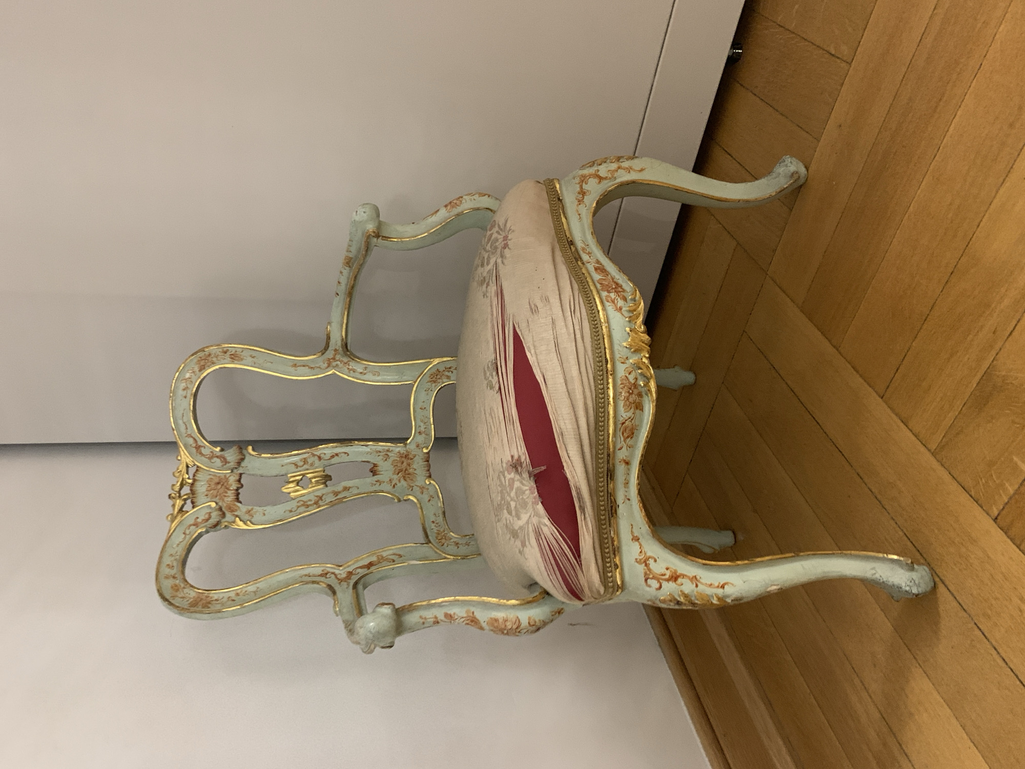 A light blue, ornate chair with gold trim and floral detailing sits on a polished wooden floor. The chair has a high, curved back and armrests. The seat of the chair is upholstered in a cream-colored fabric with a floral pattern, and there is a visible tear in the fabric revealing a red layer underneath. The chair is positioned in the corner of a room, with a white wall on the left and a light grey wall on the right. The lighting is even and appears to be artificial, casting no harsh shadows. The scene is static and appears to be an indoor setting, possibly a home or a museum display. There are no people or animals present in the image. The overall impression is one of antique furniture in a clean, well-maintained environment. The location in Poznan, Poland is not determinable from the image itself.