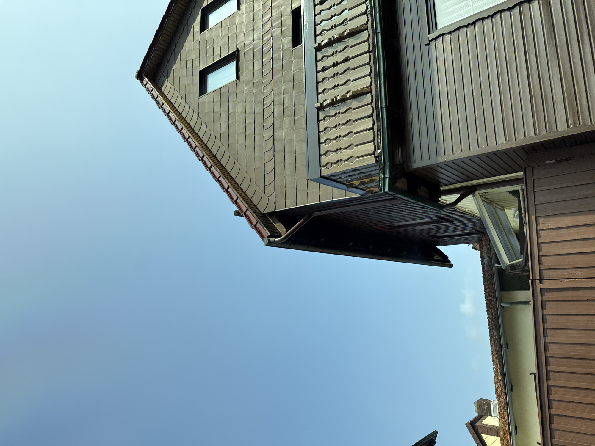 This low-angle, full-shot image captures a section of residential buildings against a clear blue sky in Bad Sachsa, Germany. The foreground features a brown, vertically-slatted fence. Behind it, a light yellow building with a red-tiled roof is partially visible, with a section of a grey downspout and a white wall.
Dominating the right and upper portion of the frame is a dark brown, wood-sided building with a grey, shingle-covered roof. The roofline is steep, and two rectangular windows with dark frames are visible on the upper level. A dark grey balcony with ornate, decorative railings extends from this building. Below the balcony, the wood siding continues down, and a green gutter runs along the edge of the roof. Further left, another building with a similar dark wood siding and a sloped roof is visible. The overall scene depicts a quiet, residential environment under a bright, cloudless sky.